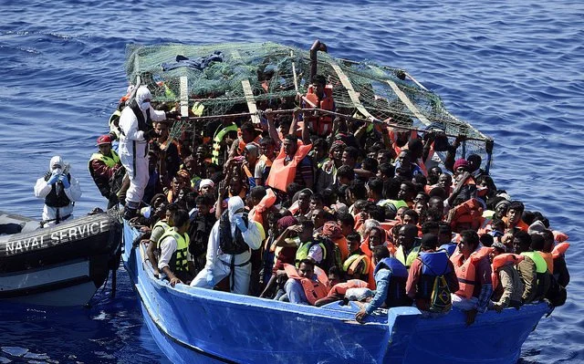 A crowded boat with many people wearing life jackets, being rescued by another boat in the ocean.