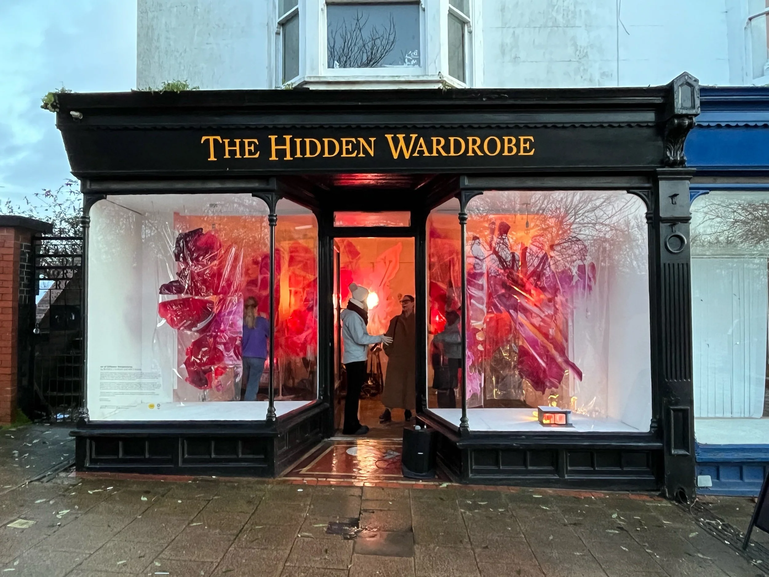 An storefront with black trim and a sign that reads 'The Hidden Wardrobe' in yellow letters. Inside, there are people viewing an art installation with red and pink hues, featuring large, abstract floral shapes covered with clear plastic.