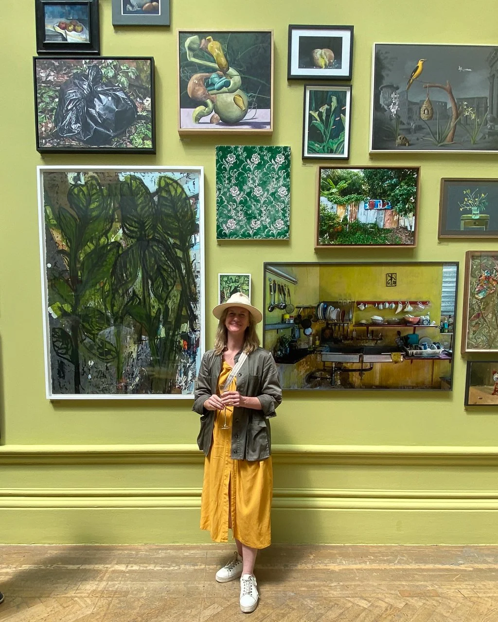 A woman wearing a yellow dress, gray jacket, white sneakers, and a wide-brimmed hat, holding a glass of wine, standing in front of a gallery wall with various colorful paintings and photographs. She is inside an art gallery or museum.