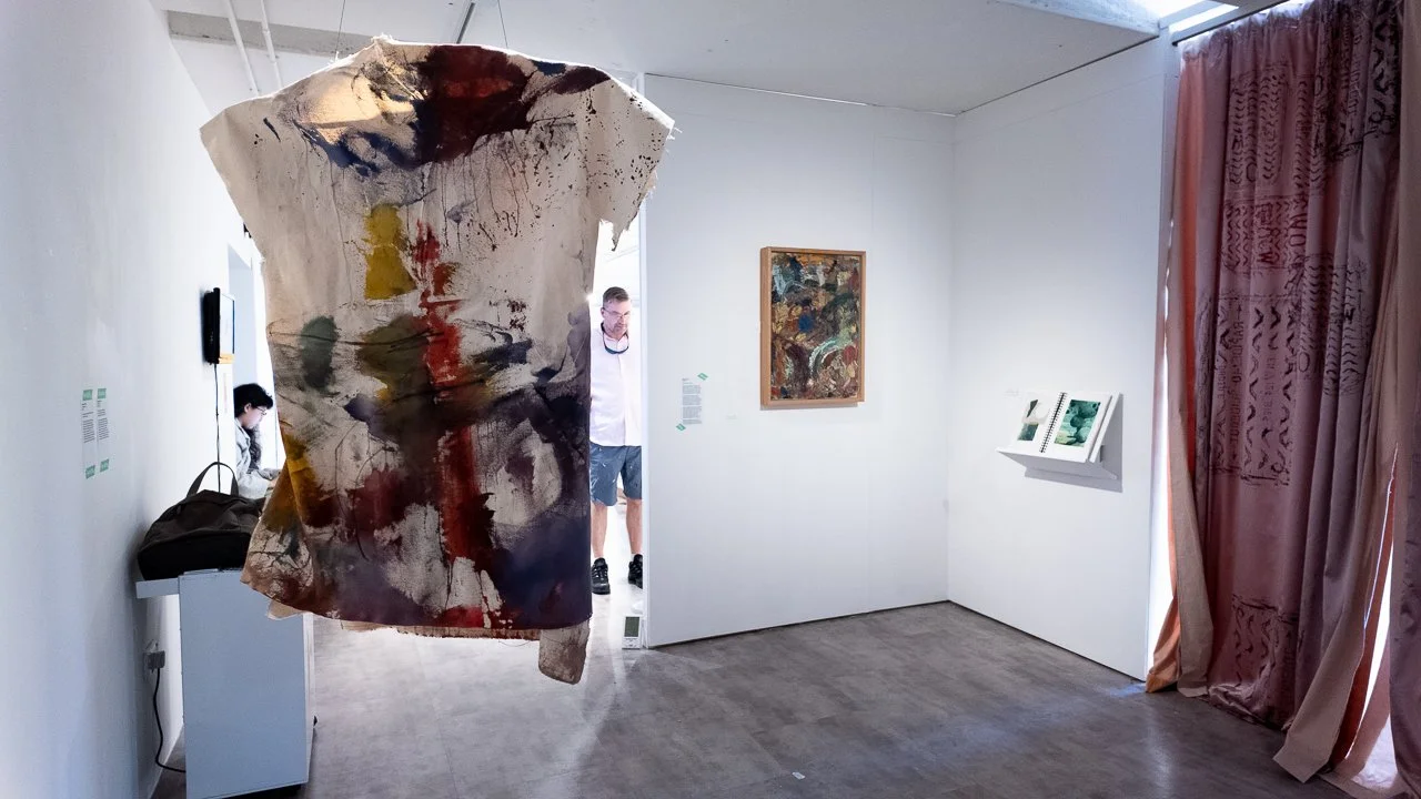 An art gallery with a large abstract painting hanging in the foreground, and a smaller framed artwork and some books on display on the wall in the background. Curtains partially cover the right side of the scene.