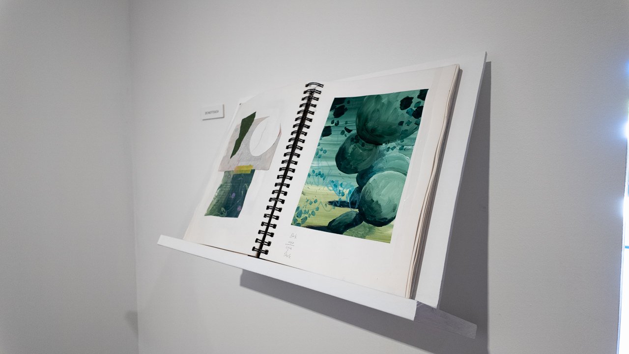 Open sketchbook on a wall displaying abstract paintings with green and blue hues.