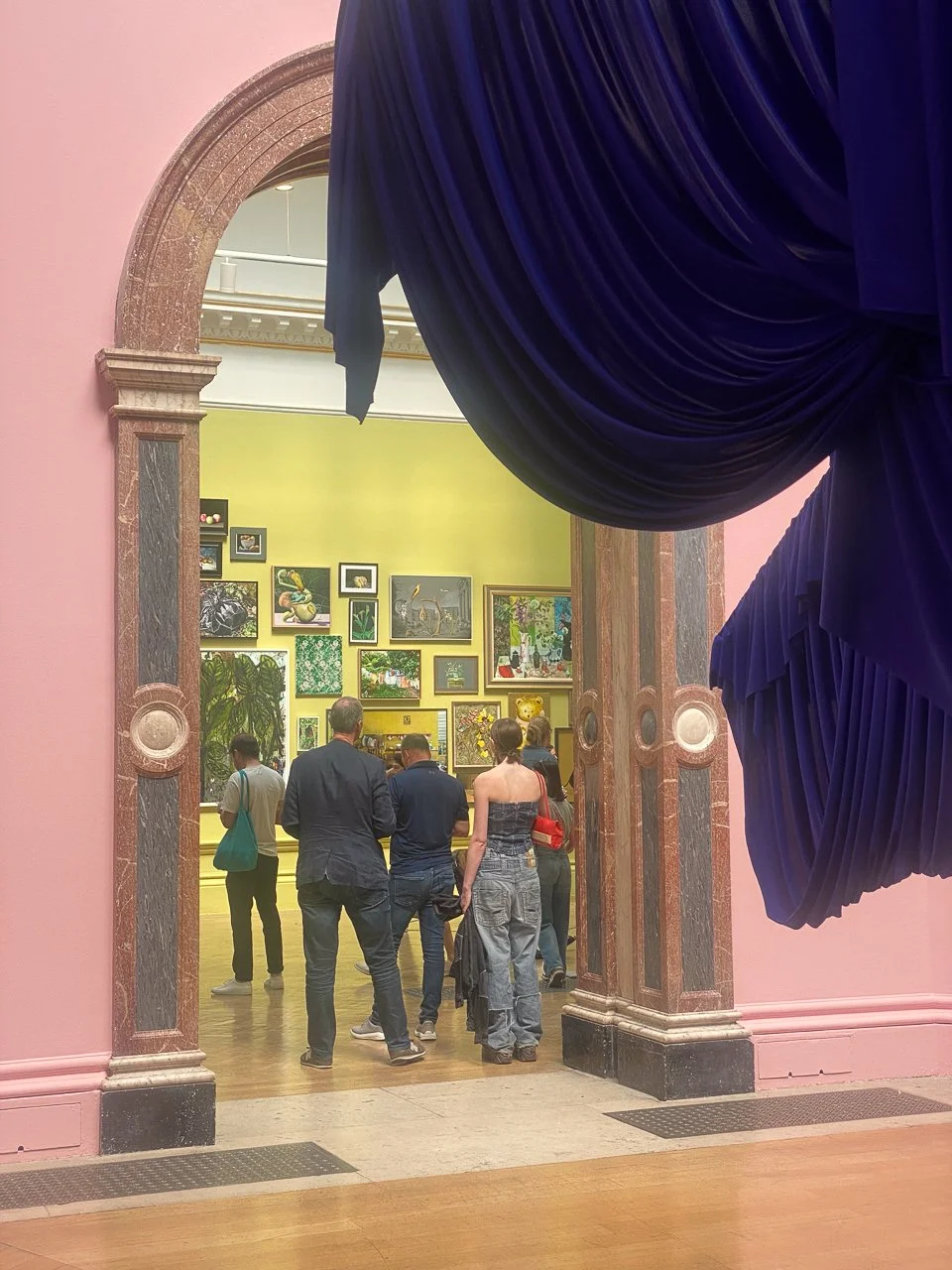 View of an art gallery through a large, arched doorway framed by pink walls and blue velvet curtains. Inside, people are observing a collection of framed artwork on a green wall.