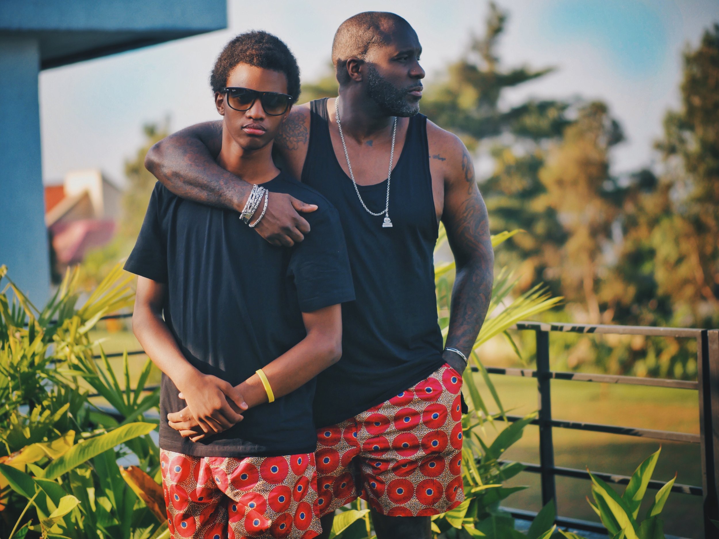  Two male-presenting Black people with matching outfits. They are both wearing black tops and red pants with a circle pattern. The person in the back has their arm around the person in front and is looking to their left. The person in front is starin
