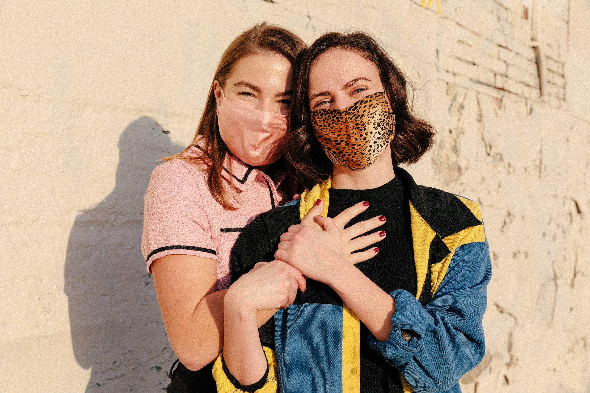  A pair of female-presenting people posing for the camera against a cream colored wall wearing face masks. The person on the left has reddish orange hair, is wearing a pink button up shirt, and is holding the person on the right. The person on the ri