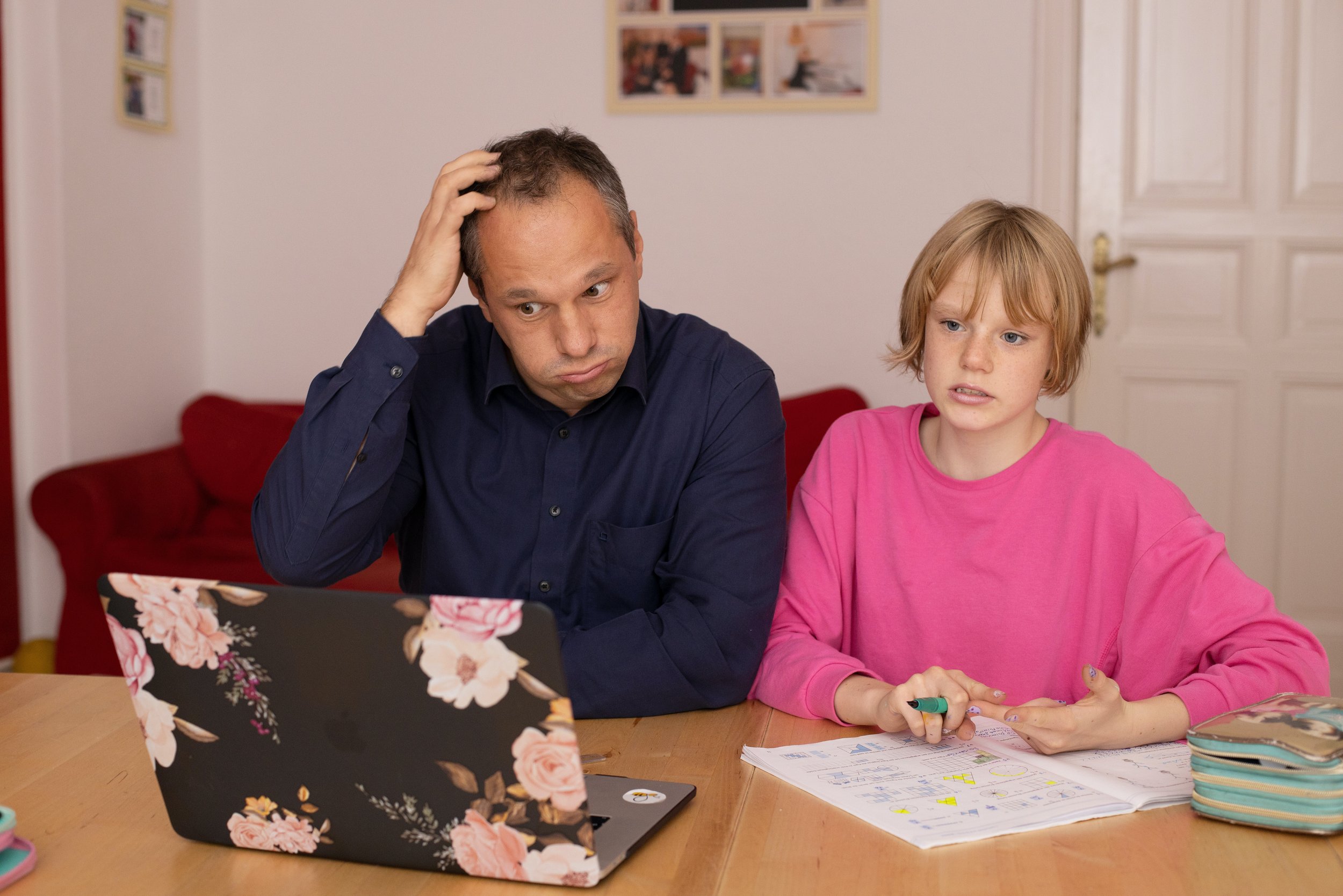  A Caucasian male-presenting father-like figure sitting next to a female-presenting younger daughter-like figure sitting next to one another facing a laptop with a black background and pink flowers. The father-like figure is scratching their head and