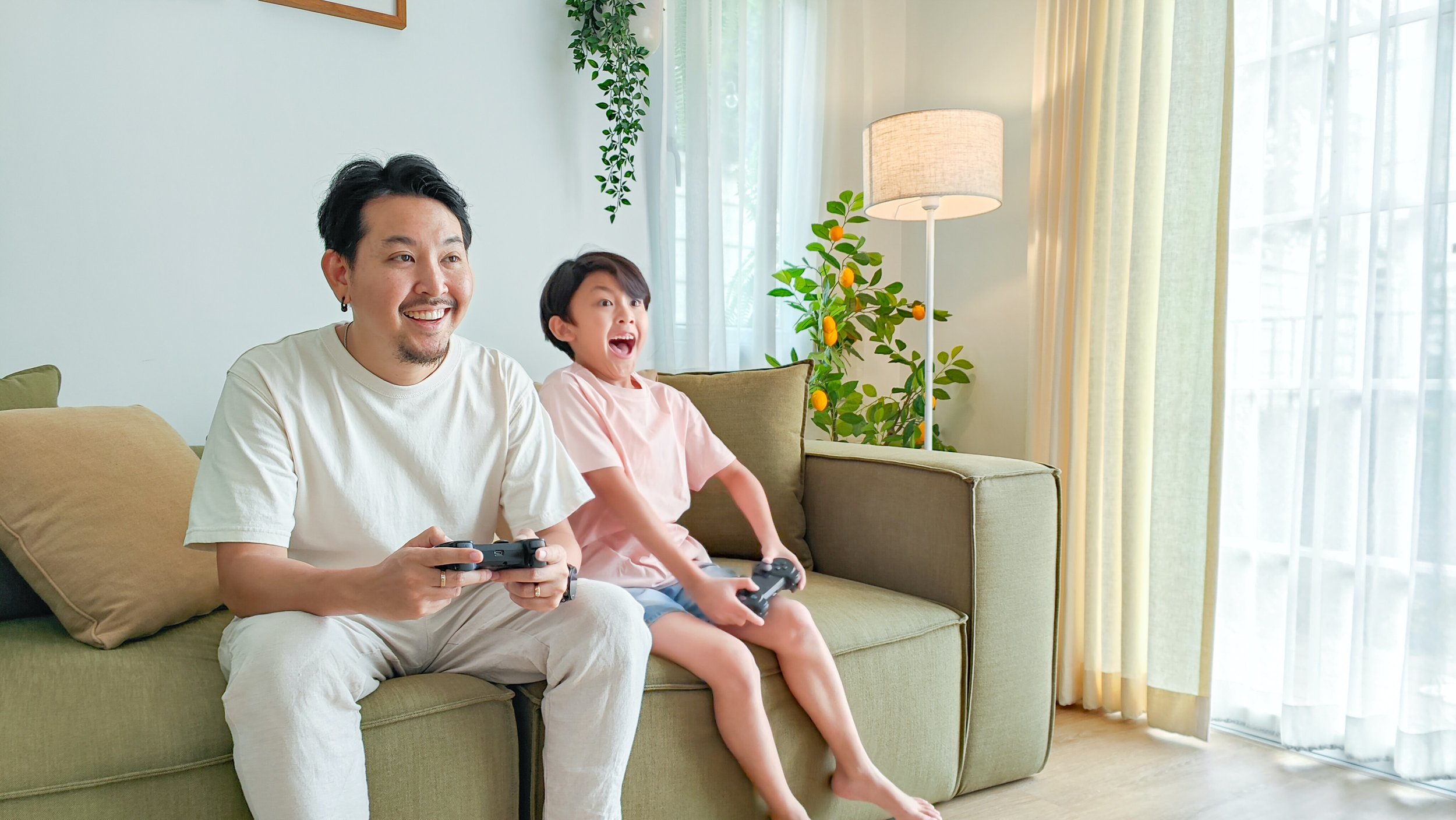 An Asian father and son presenting pair of people. They are both sitting on a green couch indoors, playing a video game with big smiles on their faces.  