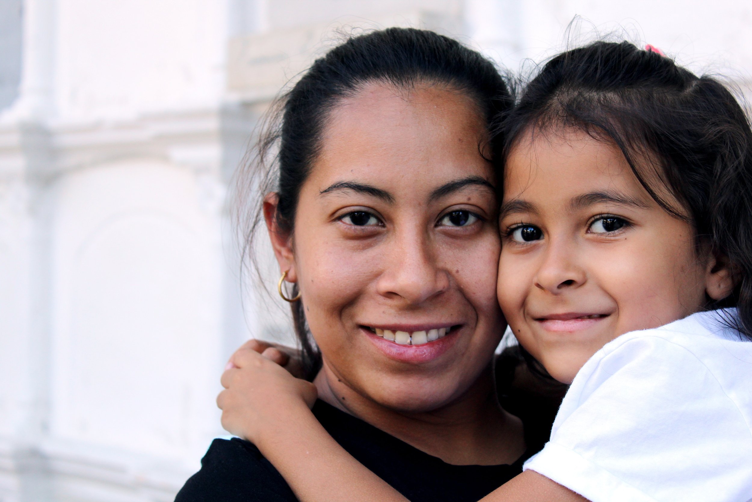  A Hispanic mother and daughter presenting pair of people. They are both facing the camera with a smile on their faces. The younger person has their arms wrapped around the mother figure.  