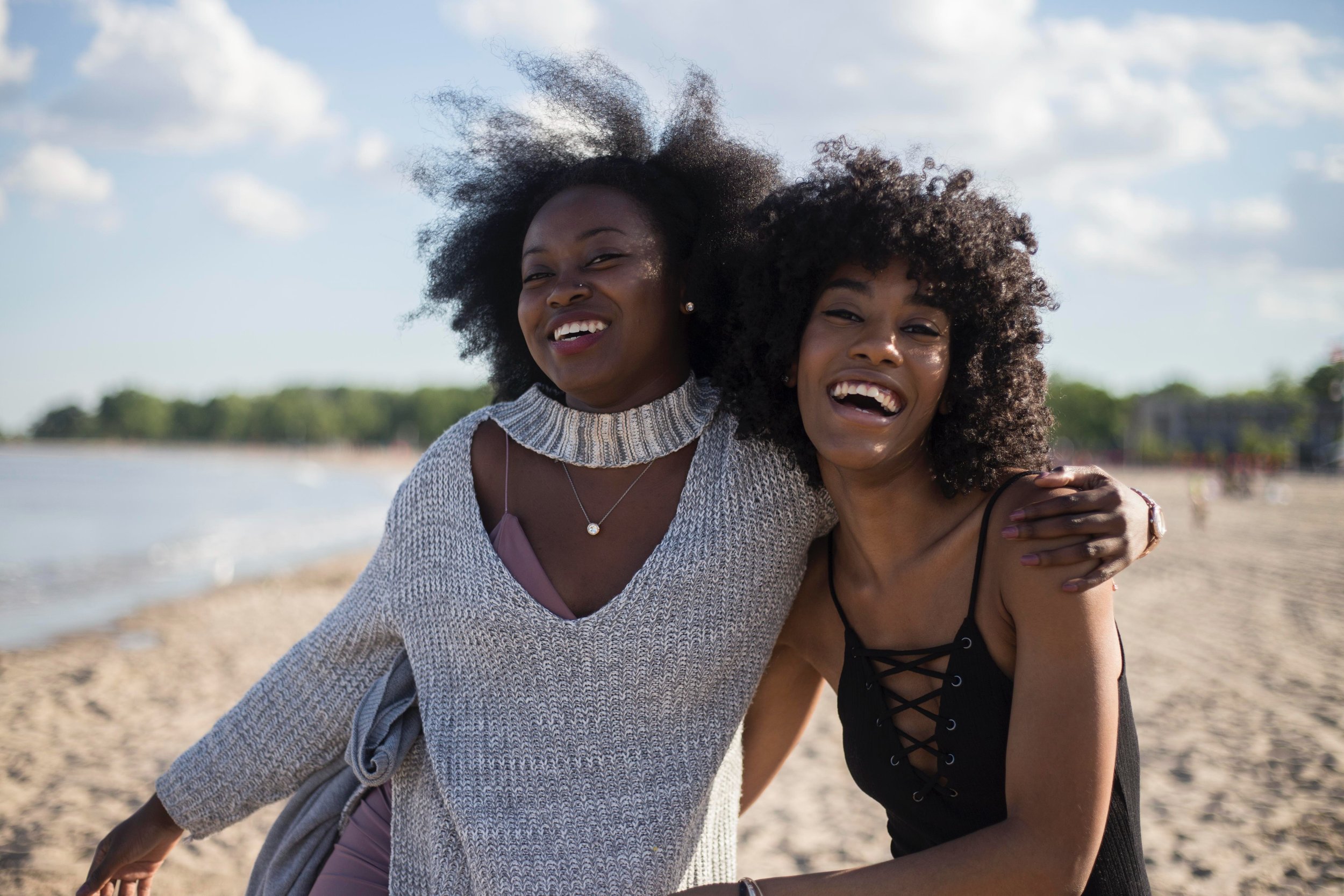  A Black pair of young female-presenting people in an outdoor setting next to a lake. They are holding each other and laughing. They both have big, curly hair. The person on the left is wearing a cutout grey sweater and the person on the right is wea