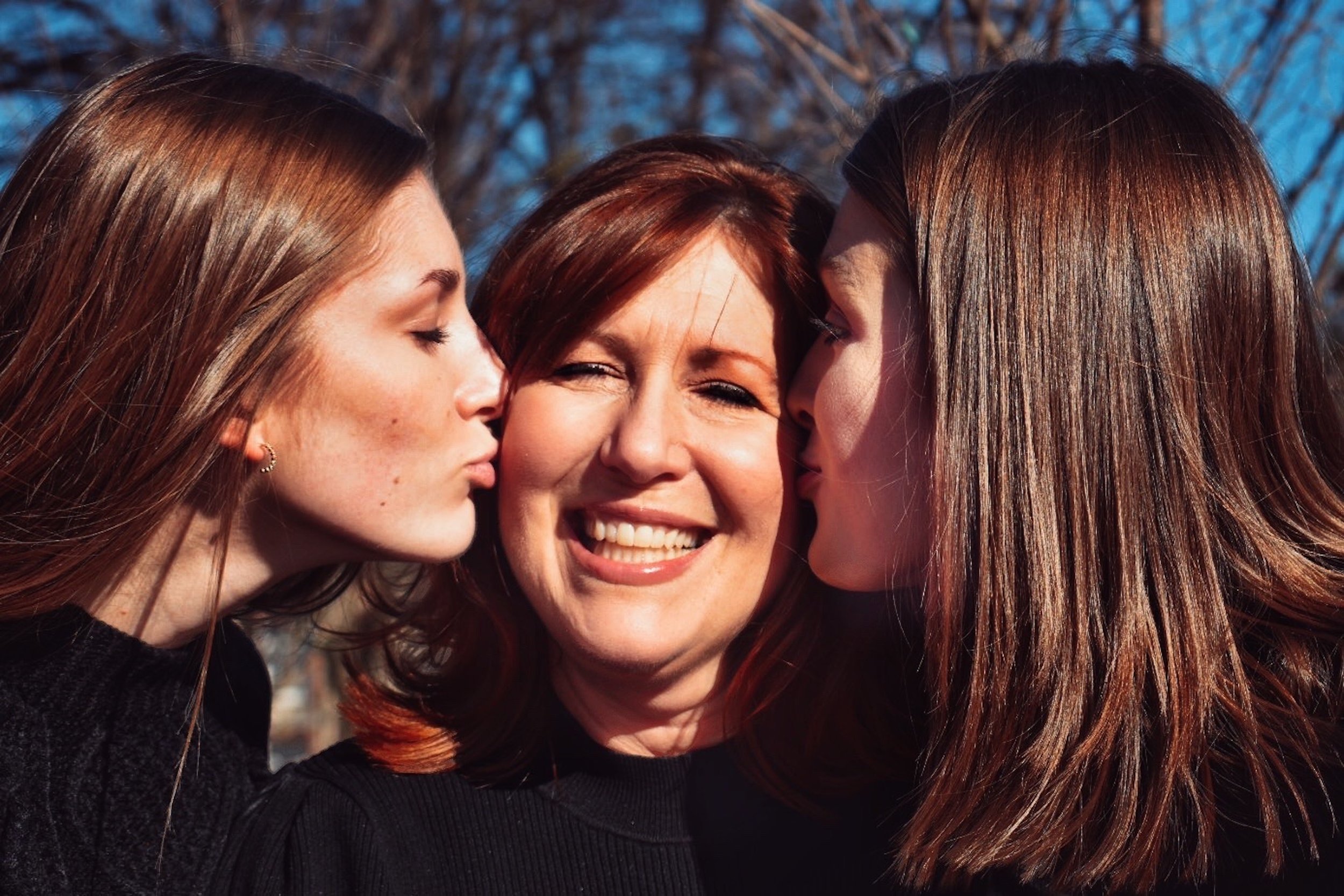  Three female-presenting Caucasian people in an outdoor setting. The person in the center is the oldest, probably the mother of the other two. On each end, the two younger people are kissing the center person on the cheek. The person in the center ha