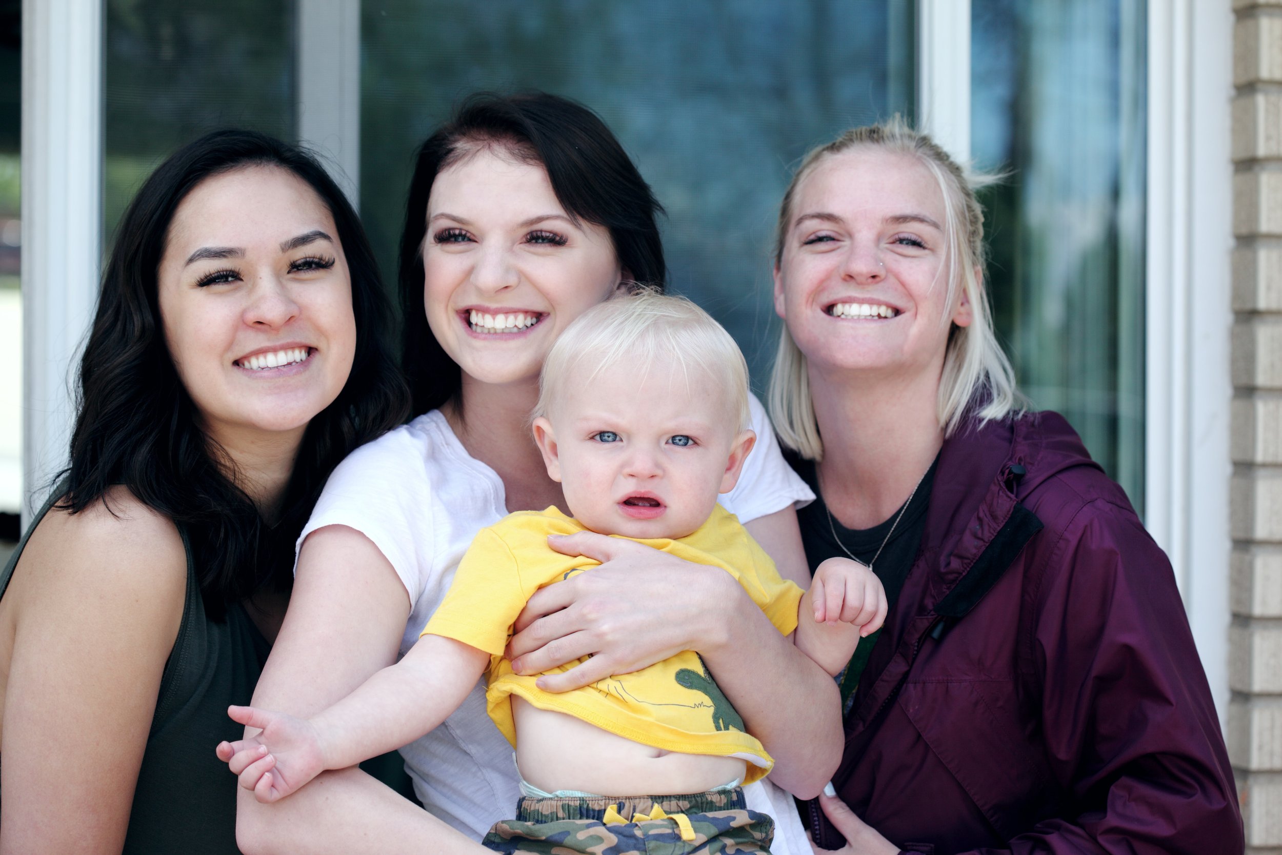  Three young female-presenting people smiling with a baby in an outdoor setting. The person on the left has black hair, the person in the center has black hair and is holding the baby. The person on the right has blonde hair. The baby is also blonde 