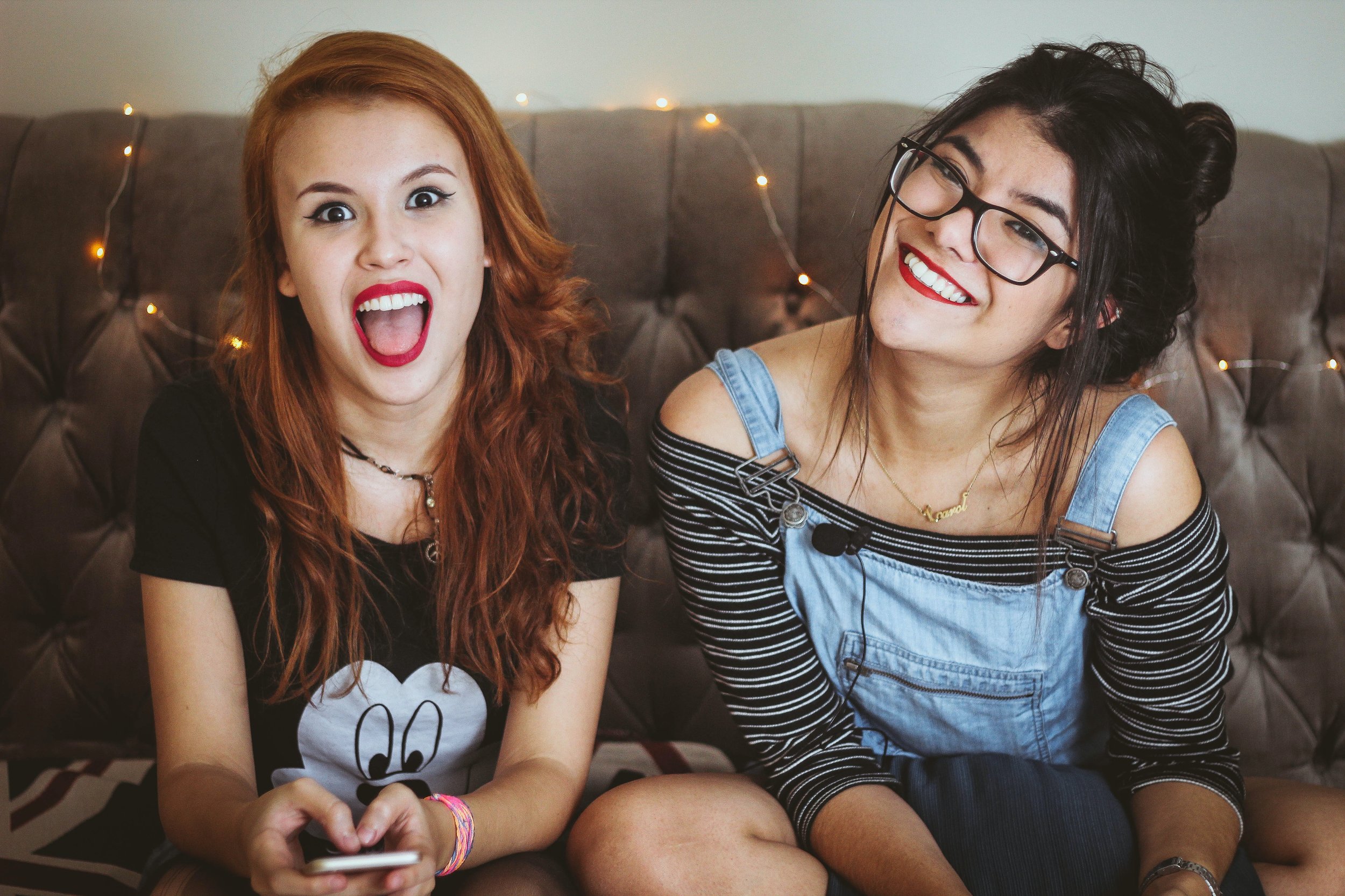  Two young female-presenting people sitting on a brown couch with lit fairy lights draped over it. The person on the left has red hair, red lipstick, and is sticking their tongue out at the camera. The person on the right has brunette hair, glasses a