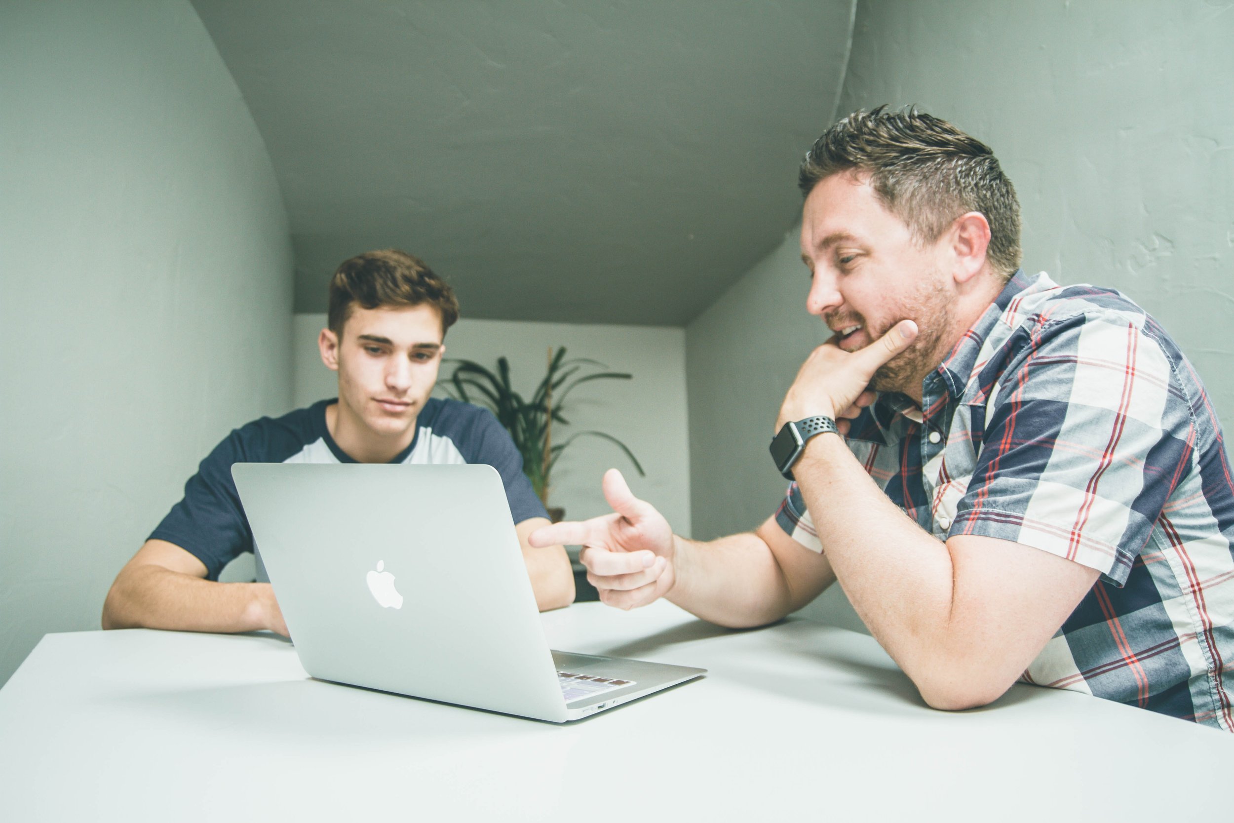  Two male-presenting people looking at a laptop in a small closet-like room. The older person is wearing a plaid shirt with grey-streaked brunette hair and a short beard. They are pointing at the laptop on the table. The younger person is wearing a b