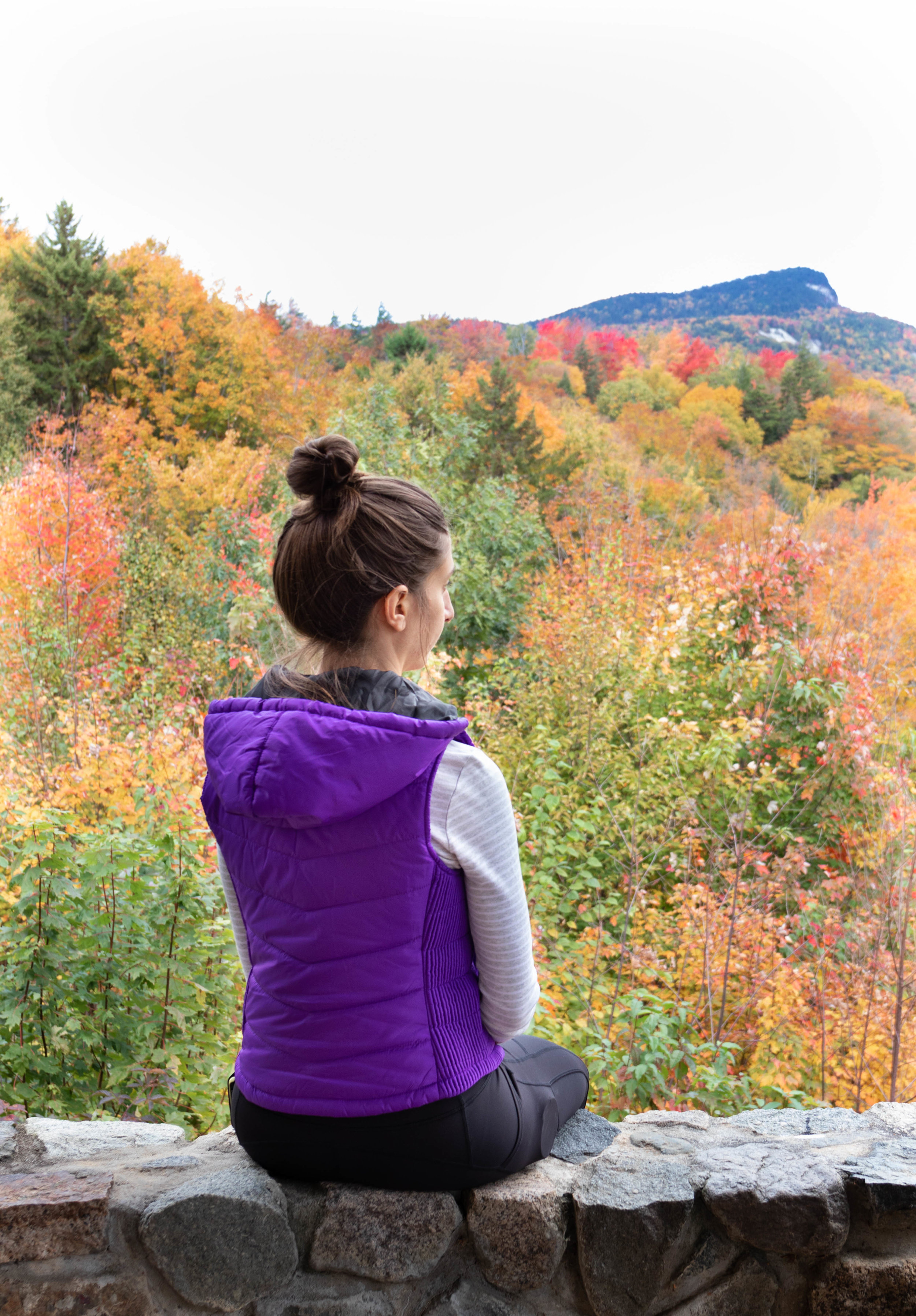 Lifestyle portrait of a woman wearing a purple vest overlooking into the fall foliage along the Kancamagus Highway in North Conway, New Hampshire by Vision Balm in Charleston, SC.