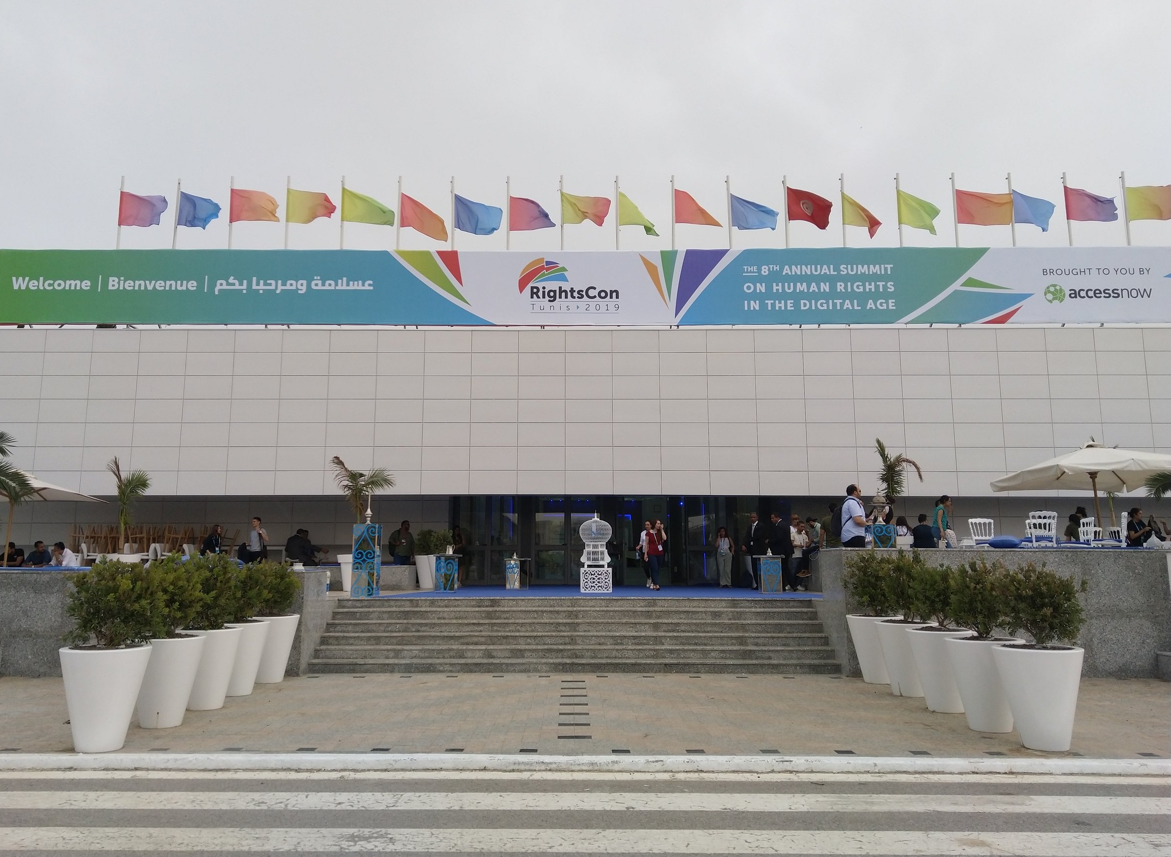 Picture showing the main entrance of the RightsCon conference venue, with a big banner over it stating “RightsCon Tunis 2019” and “8th annual summit on human rights in the digital age”. Flags on top in various colors, as well as a Tunisian flag. Pho…