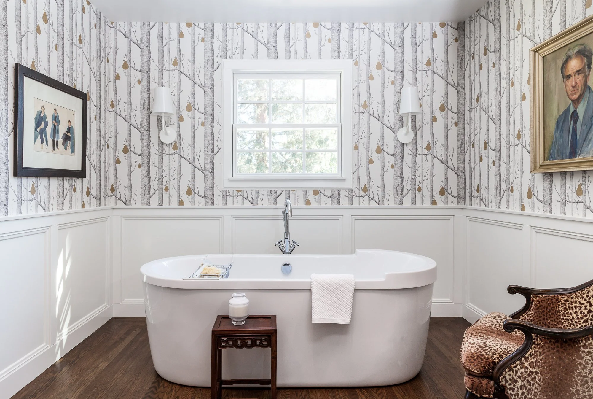 A modern bathroom with a white bathtub centered in front of a window with view of trees, surrounded by patterned wallpaper with trees and gold leaves, and decorated with framed artwork, wall-mounted lights, and a leopard print chair.