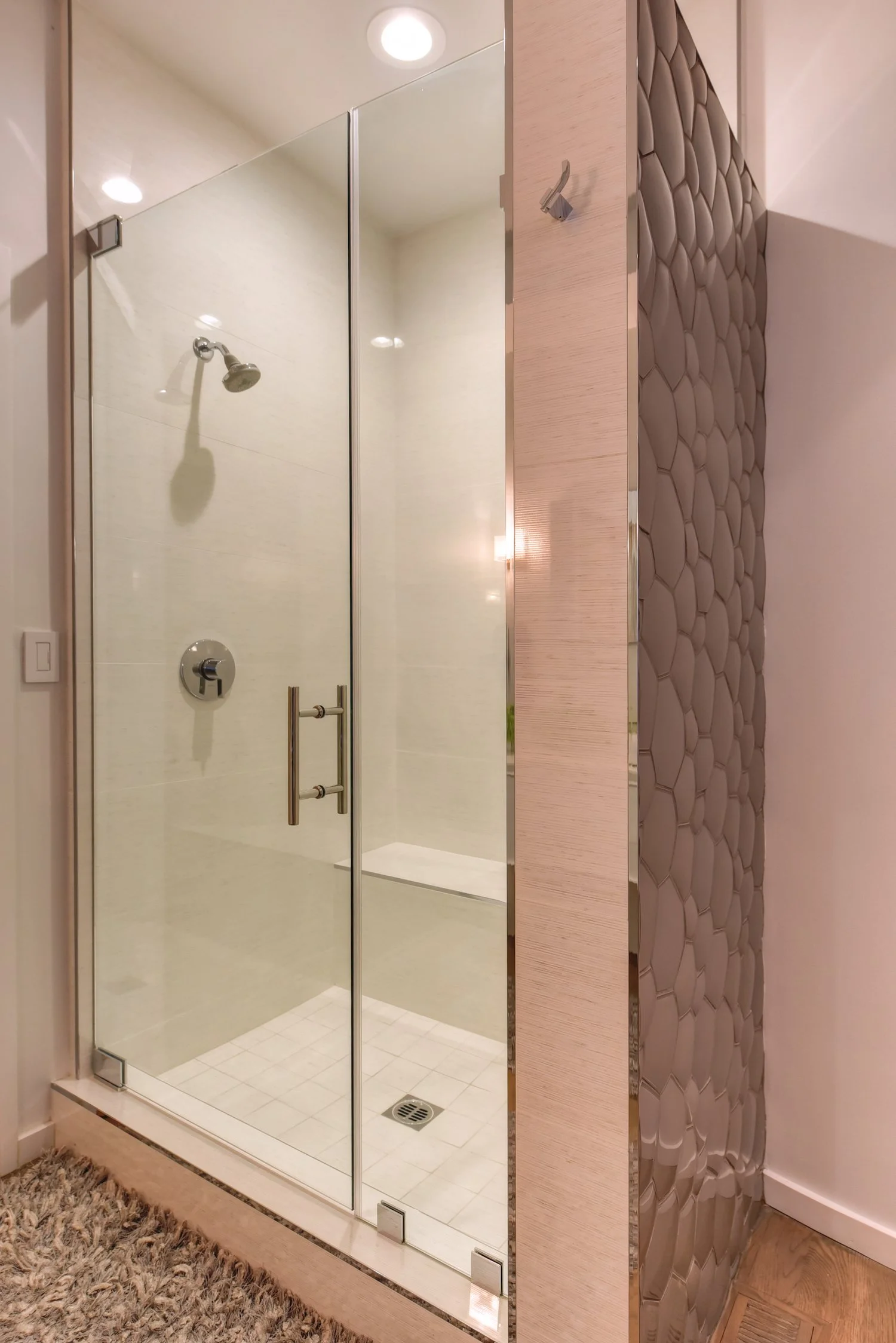 A glass enclosed shower with a metal handle on the door, a wall-mounted showerhead, and a drain on the tiled floor. The shower is situated next to a decorative wall with a textured pebble pattern.