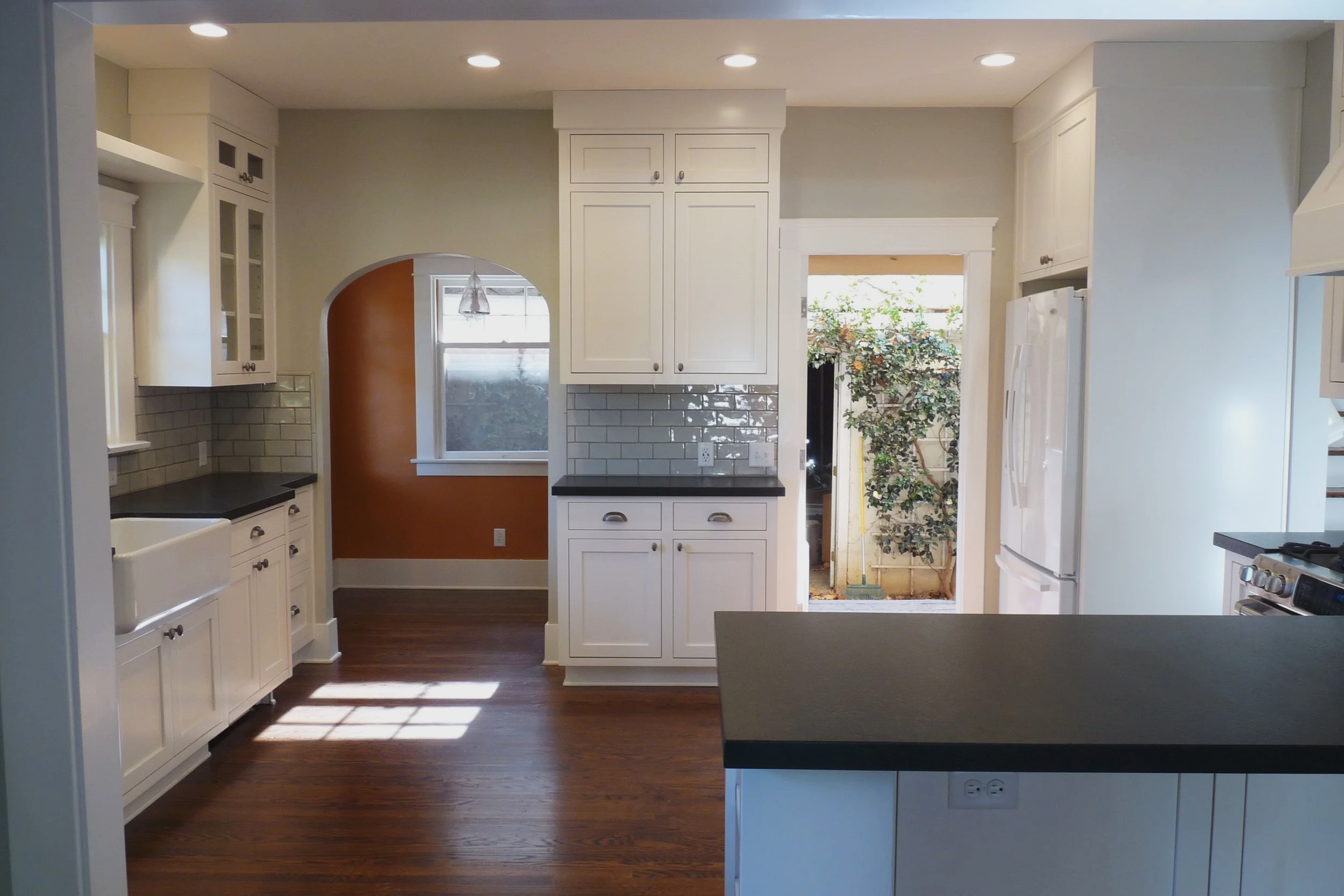 Kitchen with white cabinets, black countertops, gray subway tile backsplash, hardwood floors, and an open door leading outside to a yard with trees and plants.