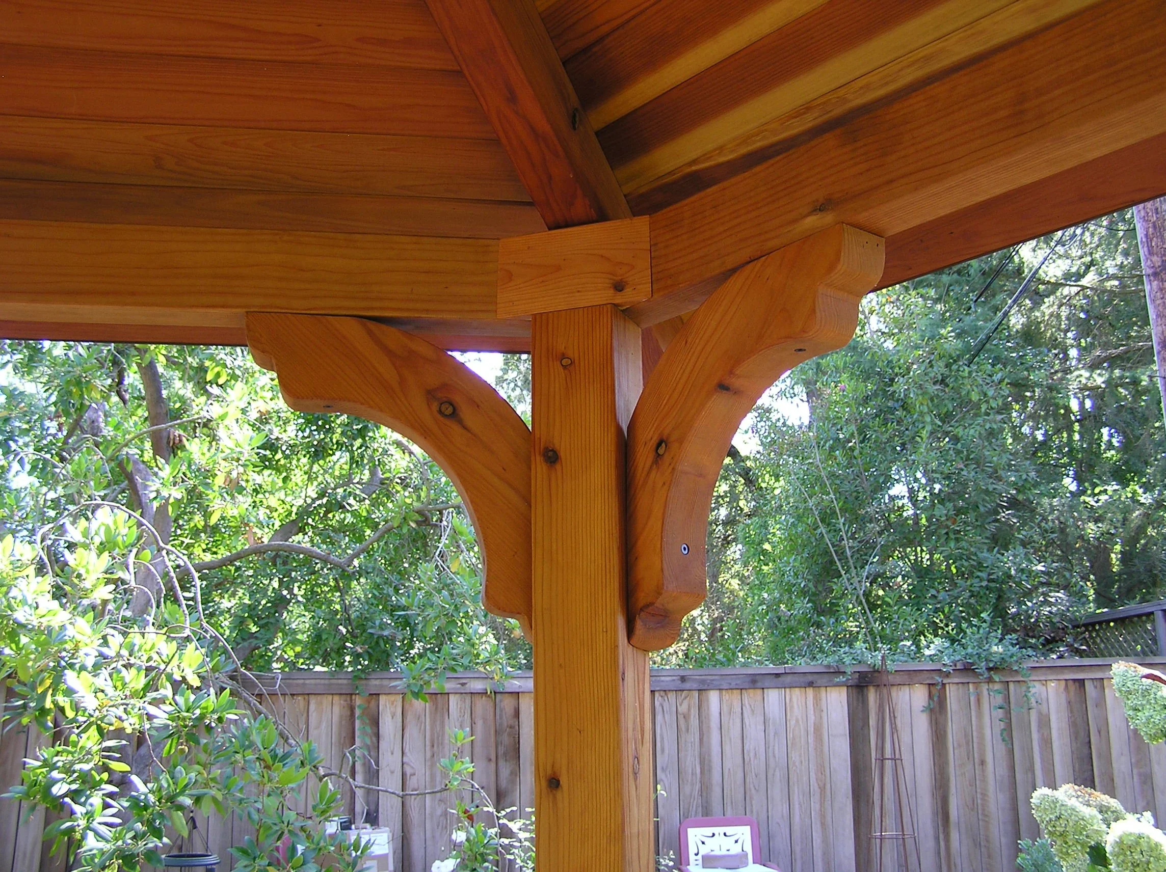 Close-up of a wooden pergola corner post and rafters with a backyard wooden fence and green trees in the background.