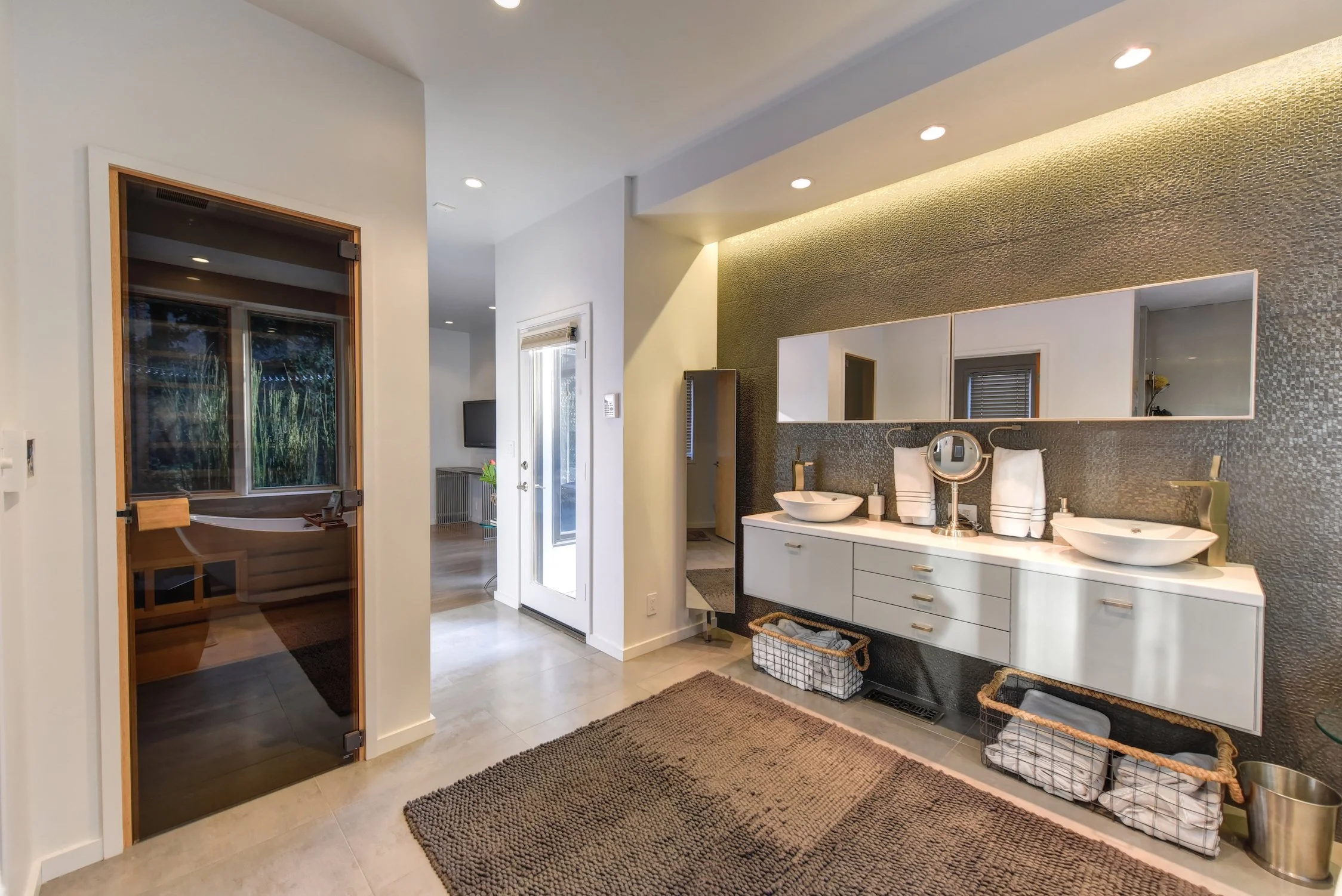 Modern bathroom with dual vessel sinks, large mirror, textured green accent wall, and woven baskets for storage.