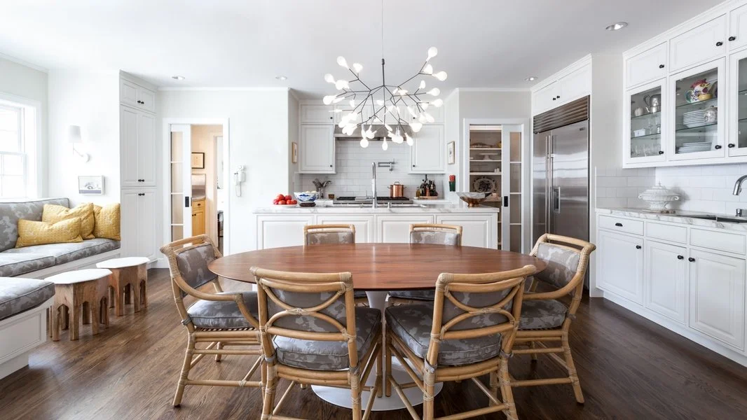 Modern kitchen with white cabinets, hardwood floors, and a round wooden dining table with eight chairs, illuminated by a starburst chandelier.