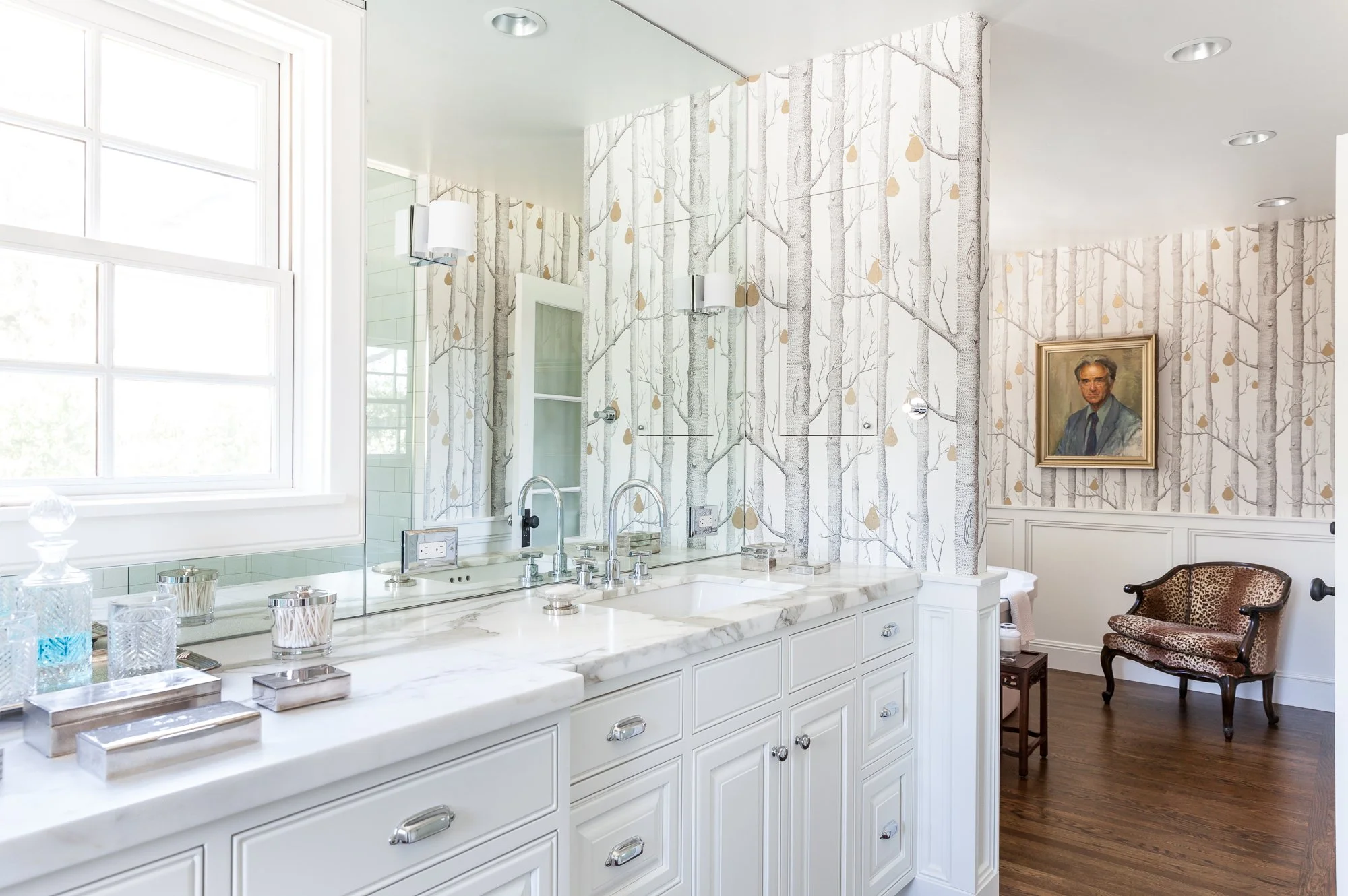 Elegant bathroom with white cabinetry, marble countertop, and wallpaper with tree branches. Mirror with sconces, window and a vintage armchair with animal print fabric. Framed portrait on the wall.