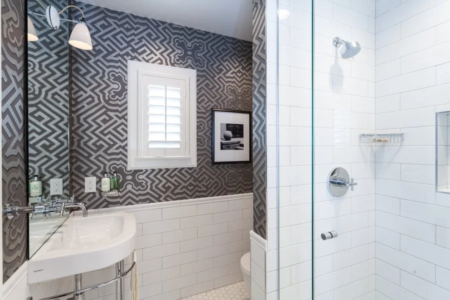 A bathroom with a geometric patterned wall, a white window with shutters, a black and white framed photo, a white oval sink, and a tiled shower area with white tiles.