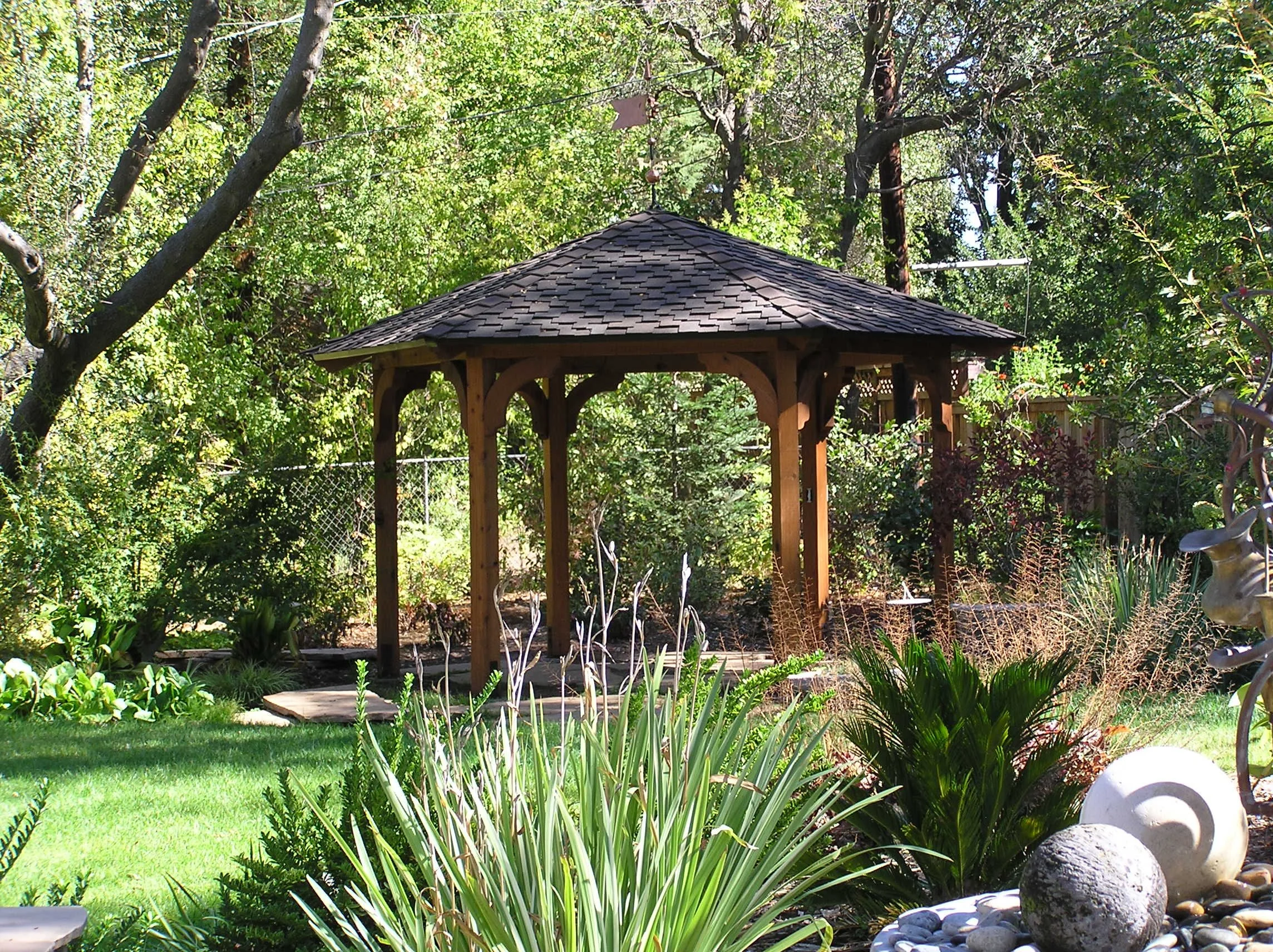 A wooden gazebo with a shingled roof in a lush garden with trees and plants.