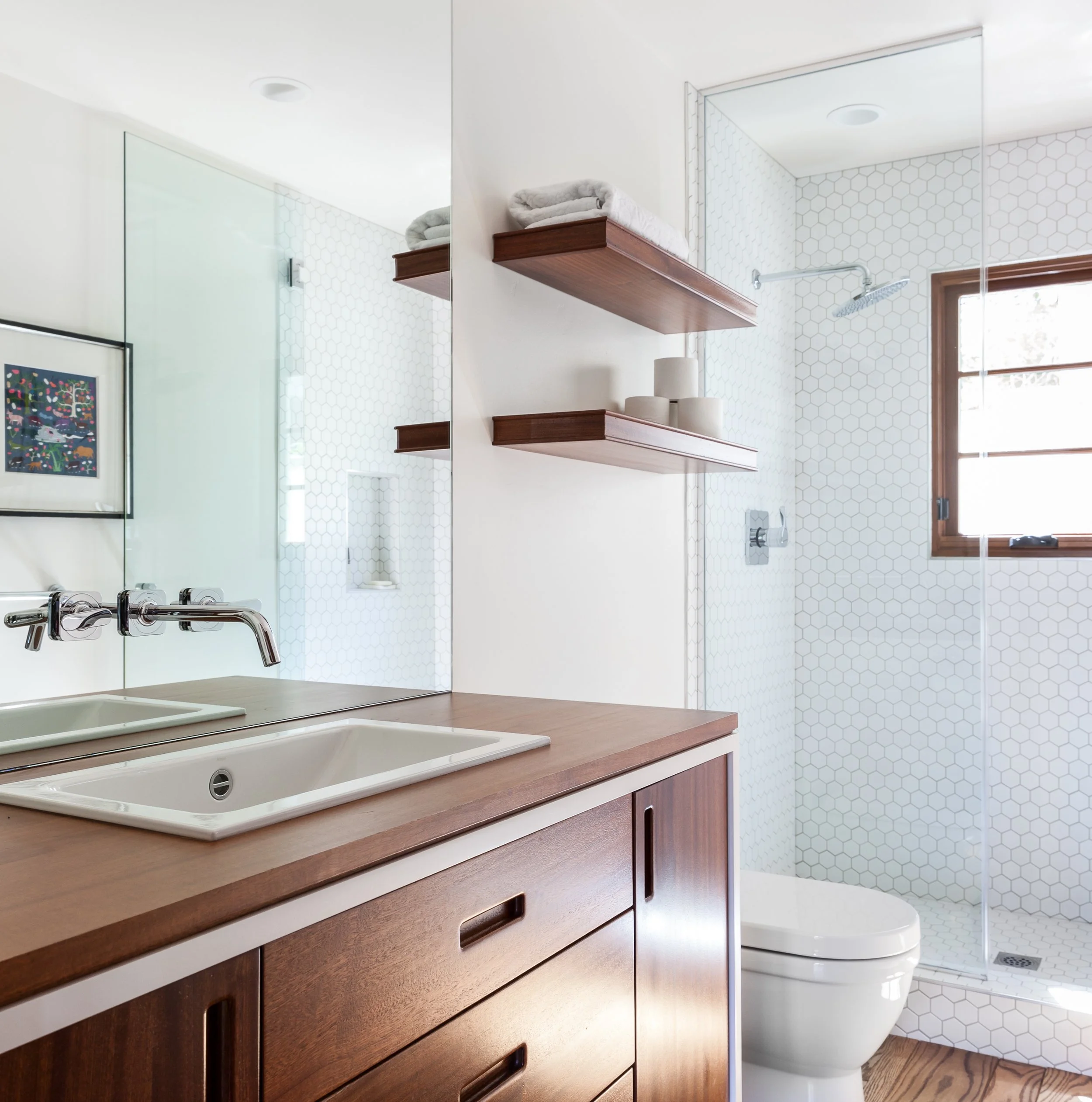 Modern bathroom with wooden vanity, white sink, large mirror, open shower with tiled walls, window, and floating wooden shelves with towels and decor.