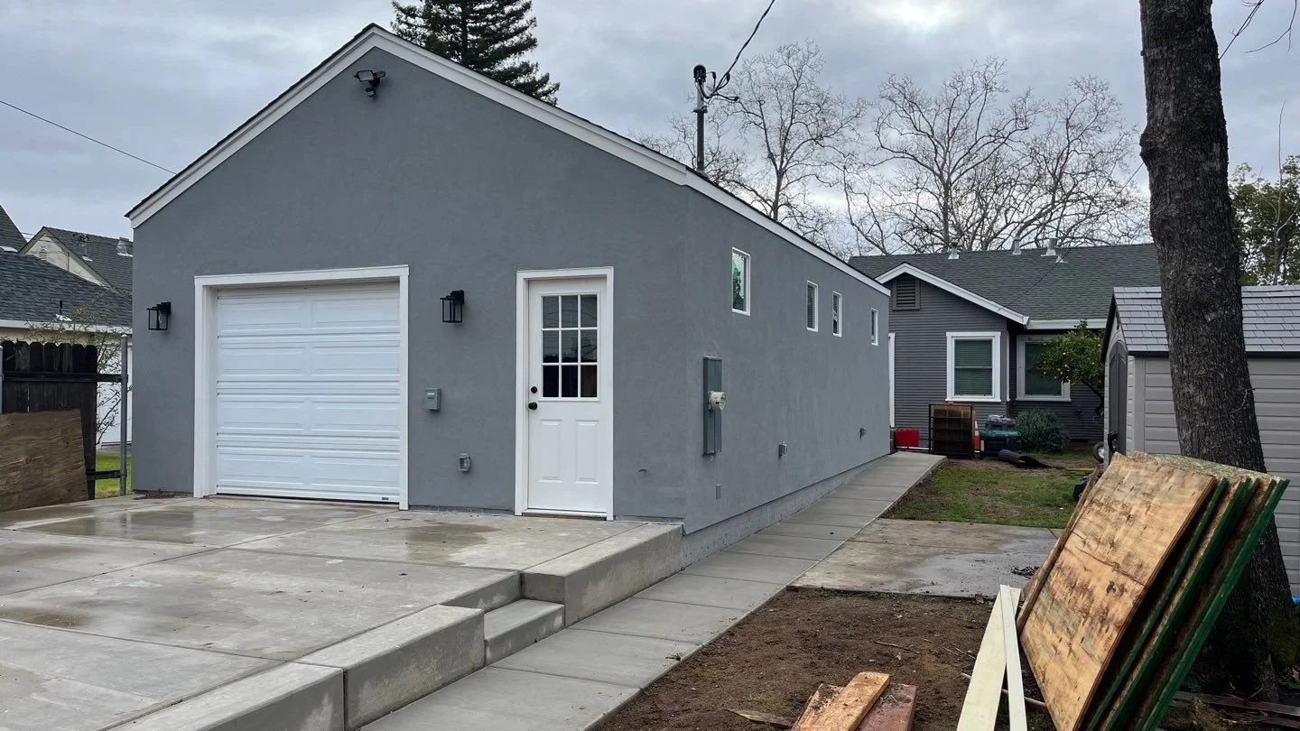 Newly constructed gray garage with white door, adjacent to small building, and sidewalk, with a large tree on the right and cloudy sky above.