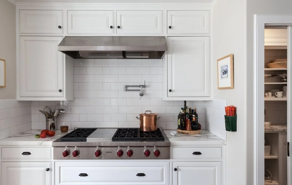 White kitchen with a stainless steel gas stove, copper pot on top, white cabinets, and minimal counter decor including tomatoes, a utensil holder, and bottles of oil or vinegar.