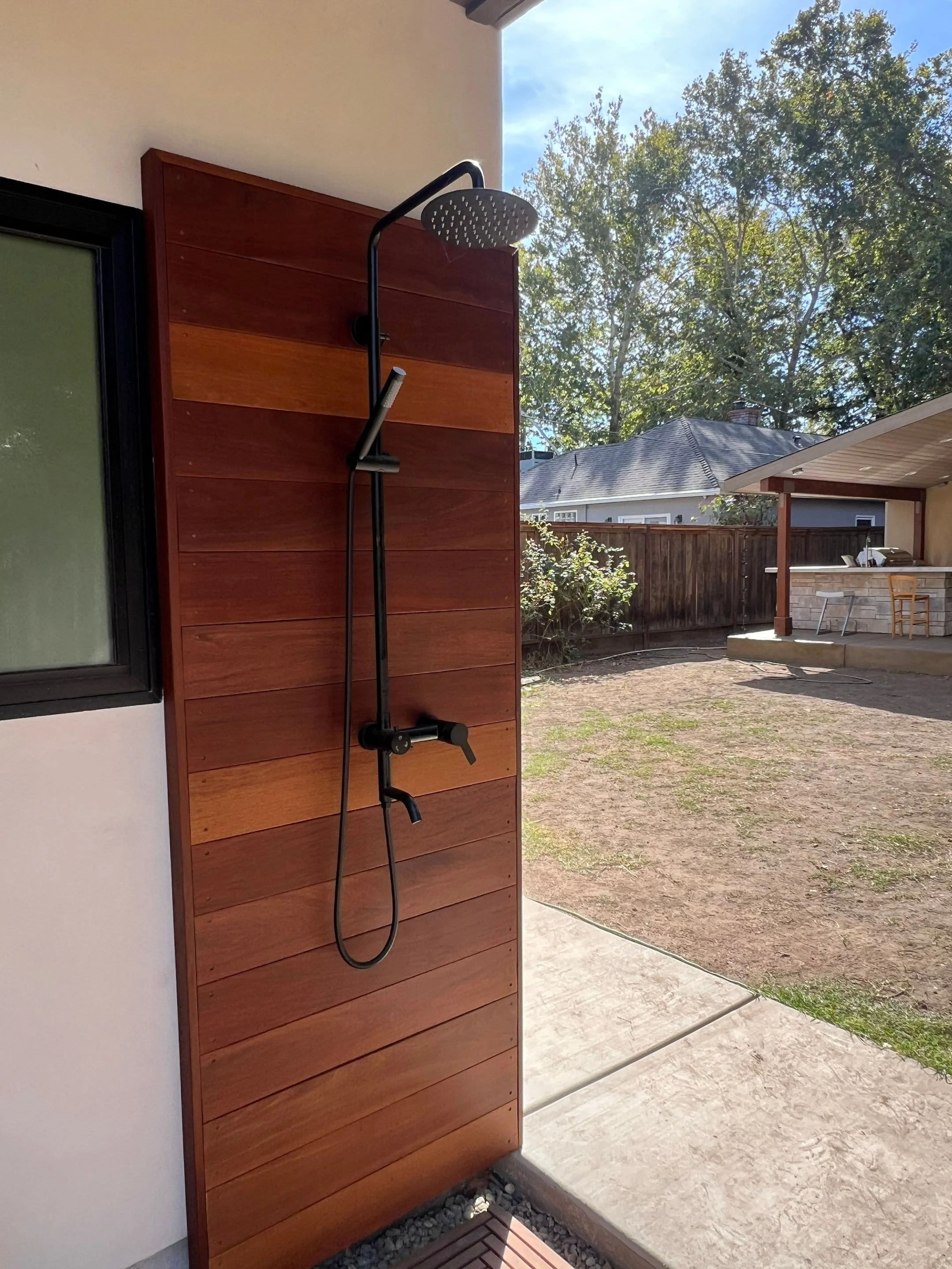 Outdoor shower with a black showerhead and control handle mounted on a vertical wooden panel outside a house.