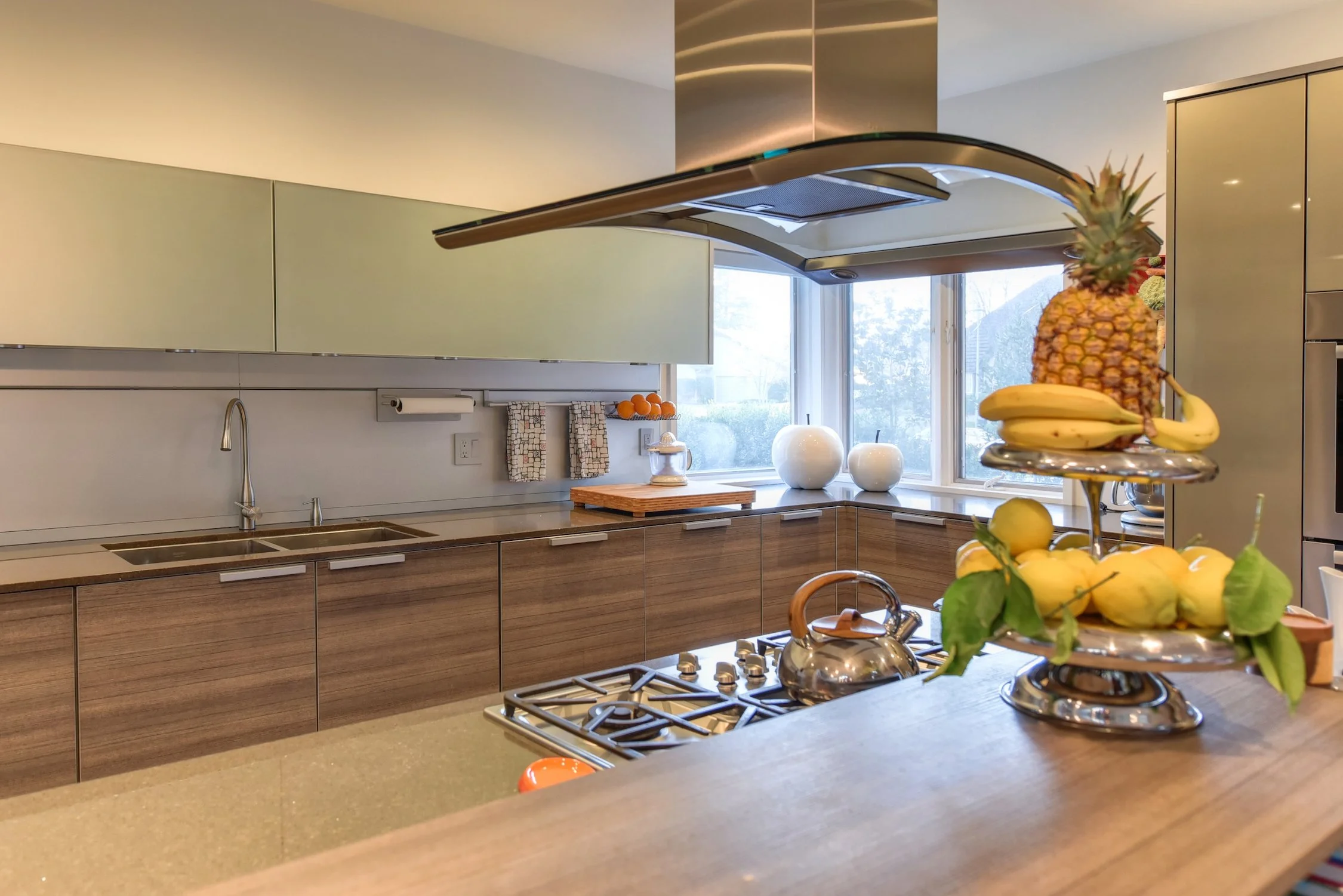 Modern kitchen with wooden lower cabinets, pastel green upper cabinets, stainless steel appliances, a stove with a teapot, a fruit stand with bananas, pineapple, lemons, and limes, and a window with a view of trees outside.