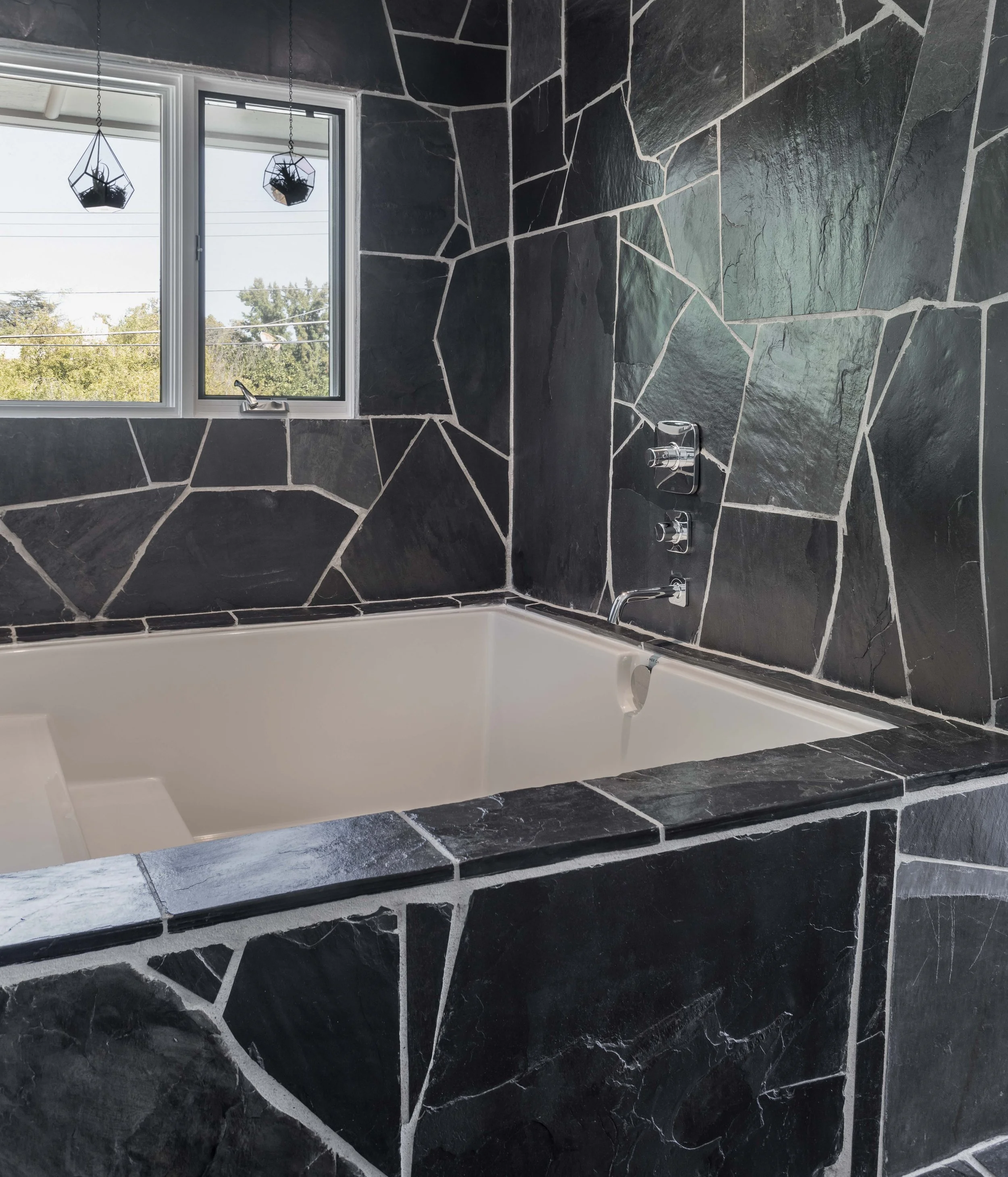 Bathroom with black slate tile walls surrounding a white bathtub and a window with two hanging plant holders.