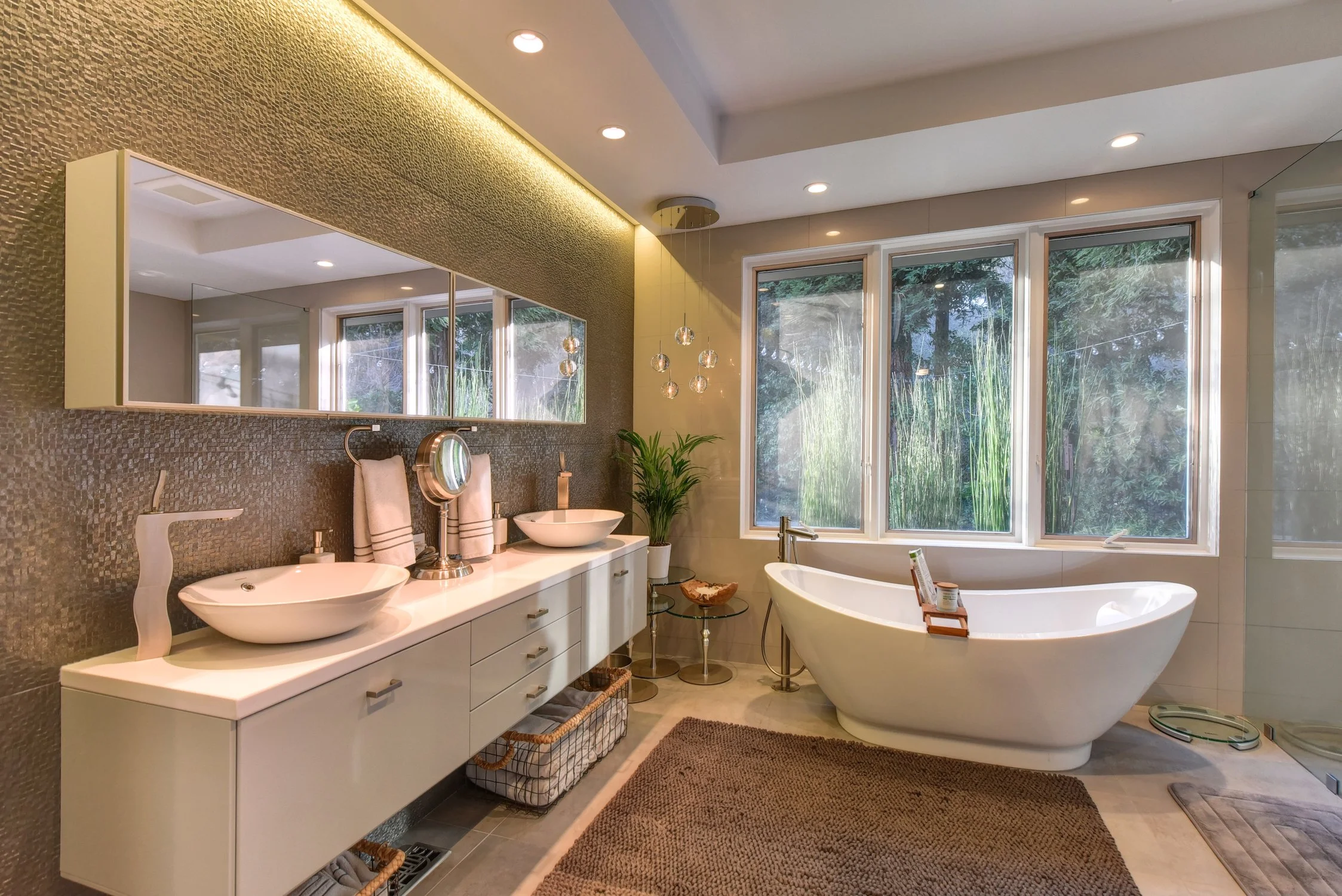 Modern bathroom with double sinks, large mirror, clawfoot bathtub by window, and shower area, decorated with a potted plant and natural light