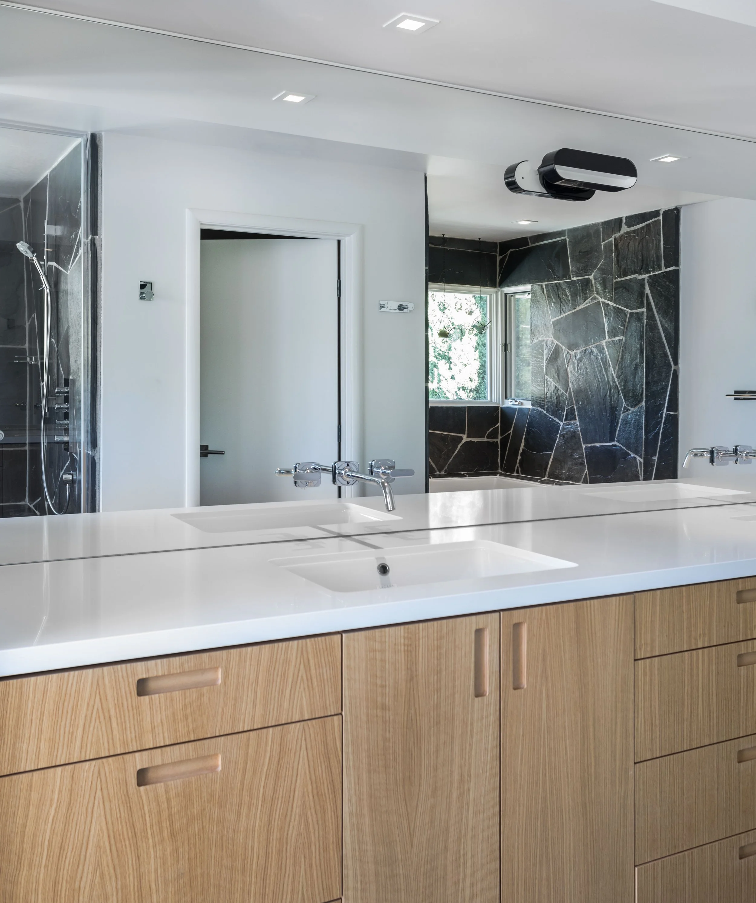 Modern bathroom with white countertop, wooden cabinetry, black stone wall, and a window with greenery outside.