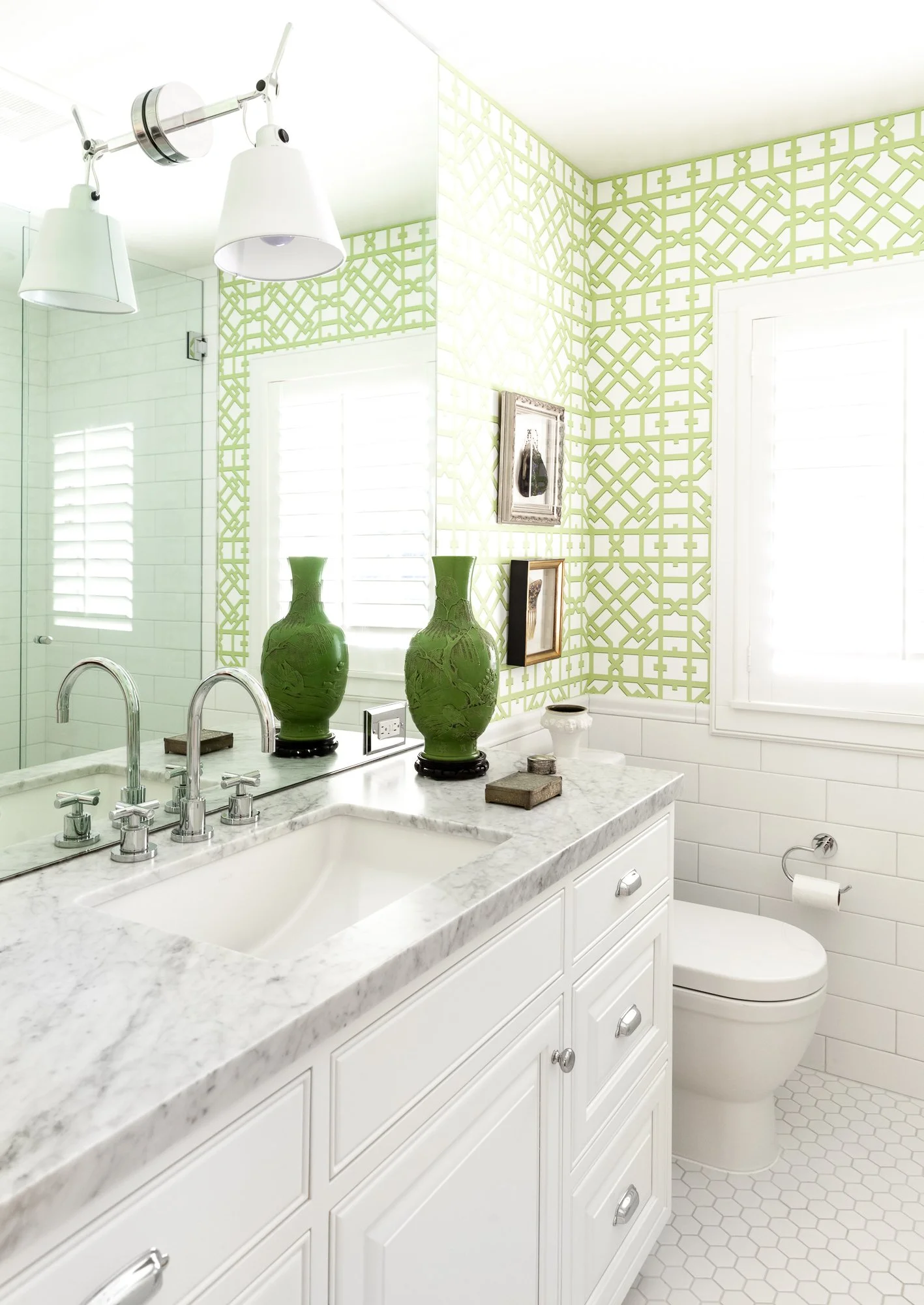 Bright bathroom with white cabinetry, marble countertop, green vases, wall mirrors, and a window with white shutters.