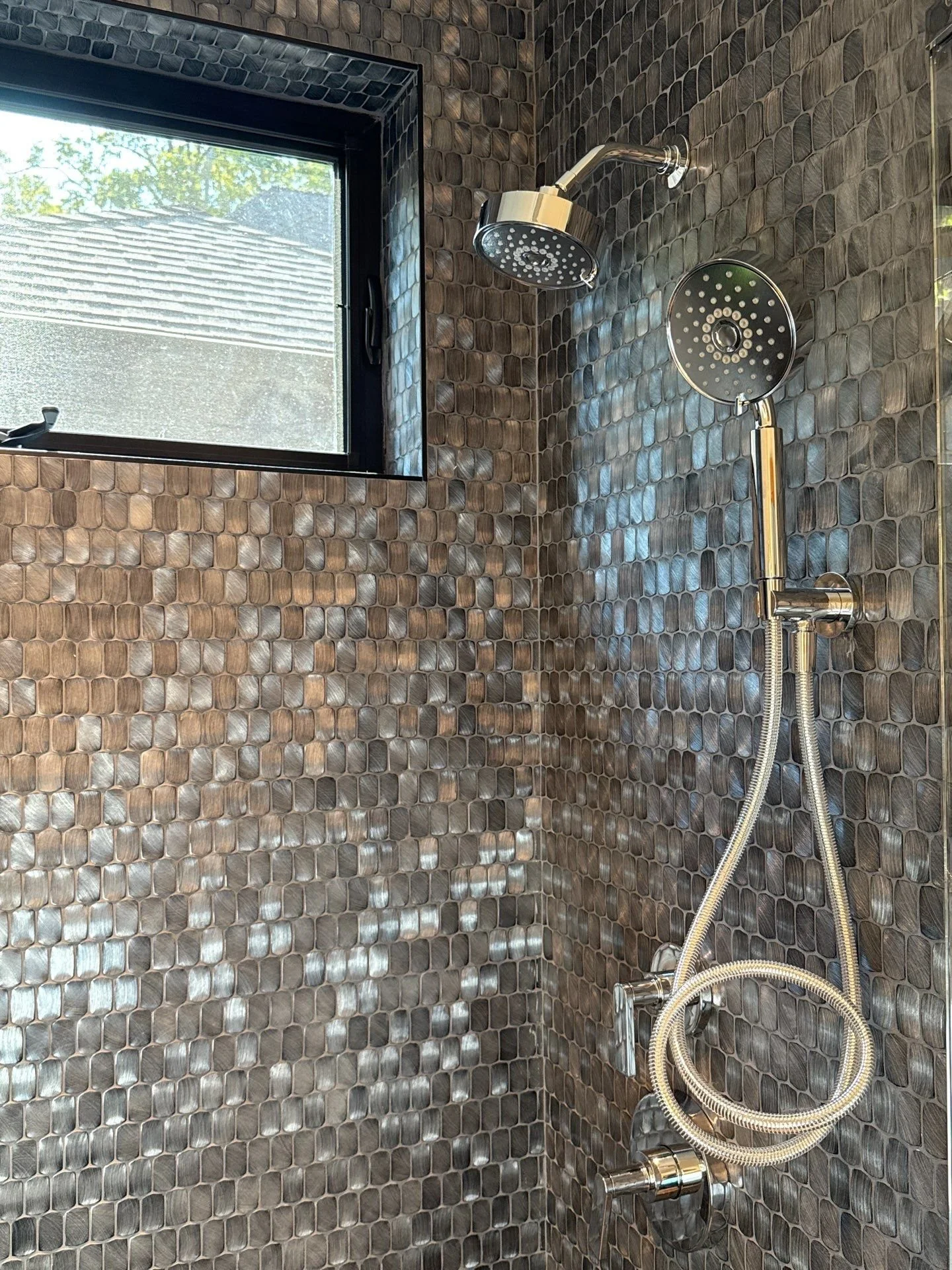A shower area with two chrome shower heads on a wall covered in small, round, brown tiles. There is a small window with a black frame above the shower, showing a view of trees and a building roof outside.