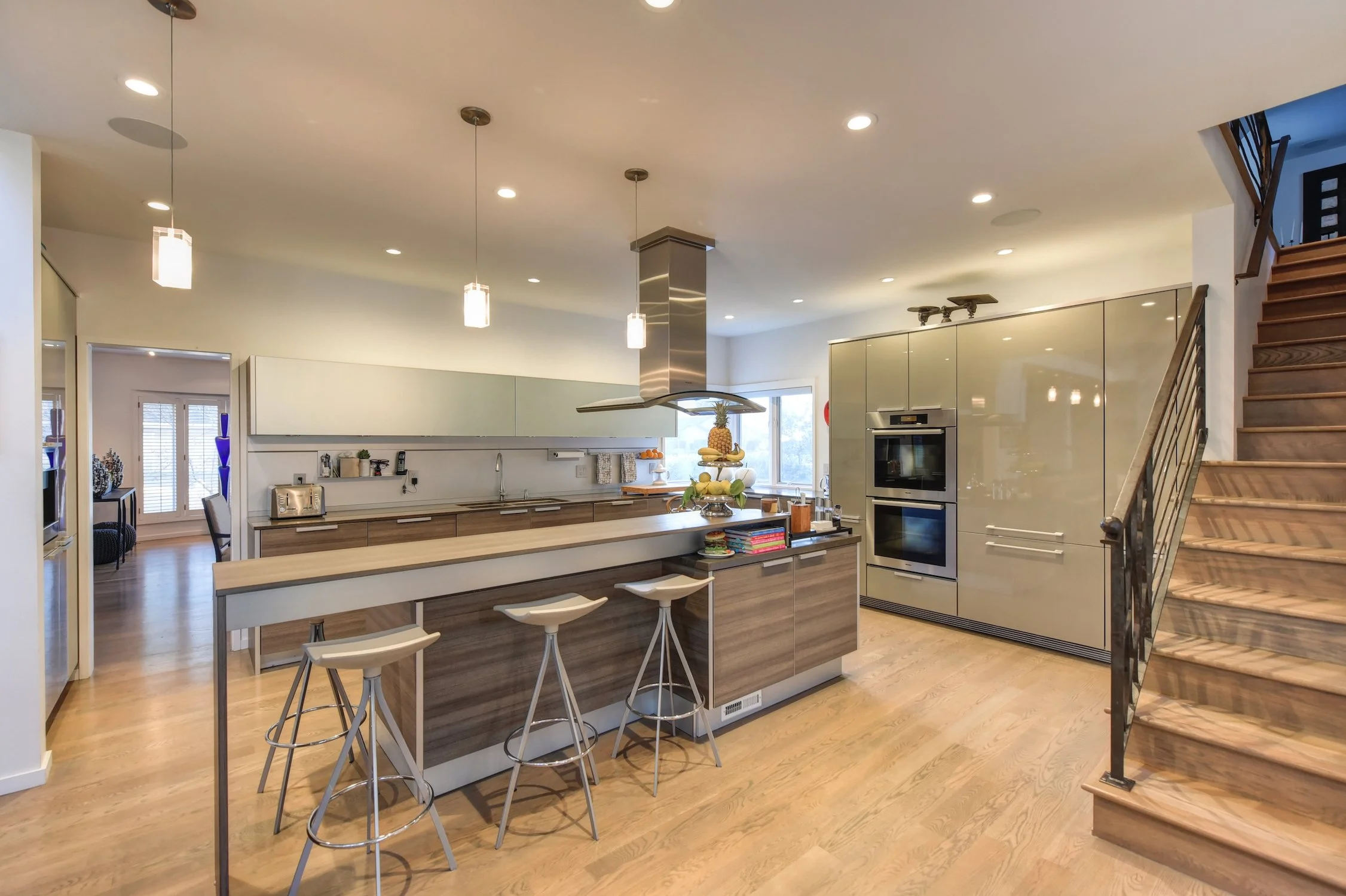 Modern kitchen with beige and wooden cabinets, a large island with bar stools, stainless steel appliances, and a staircase to the right.