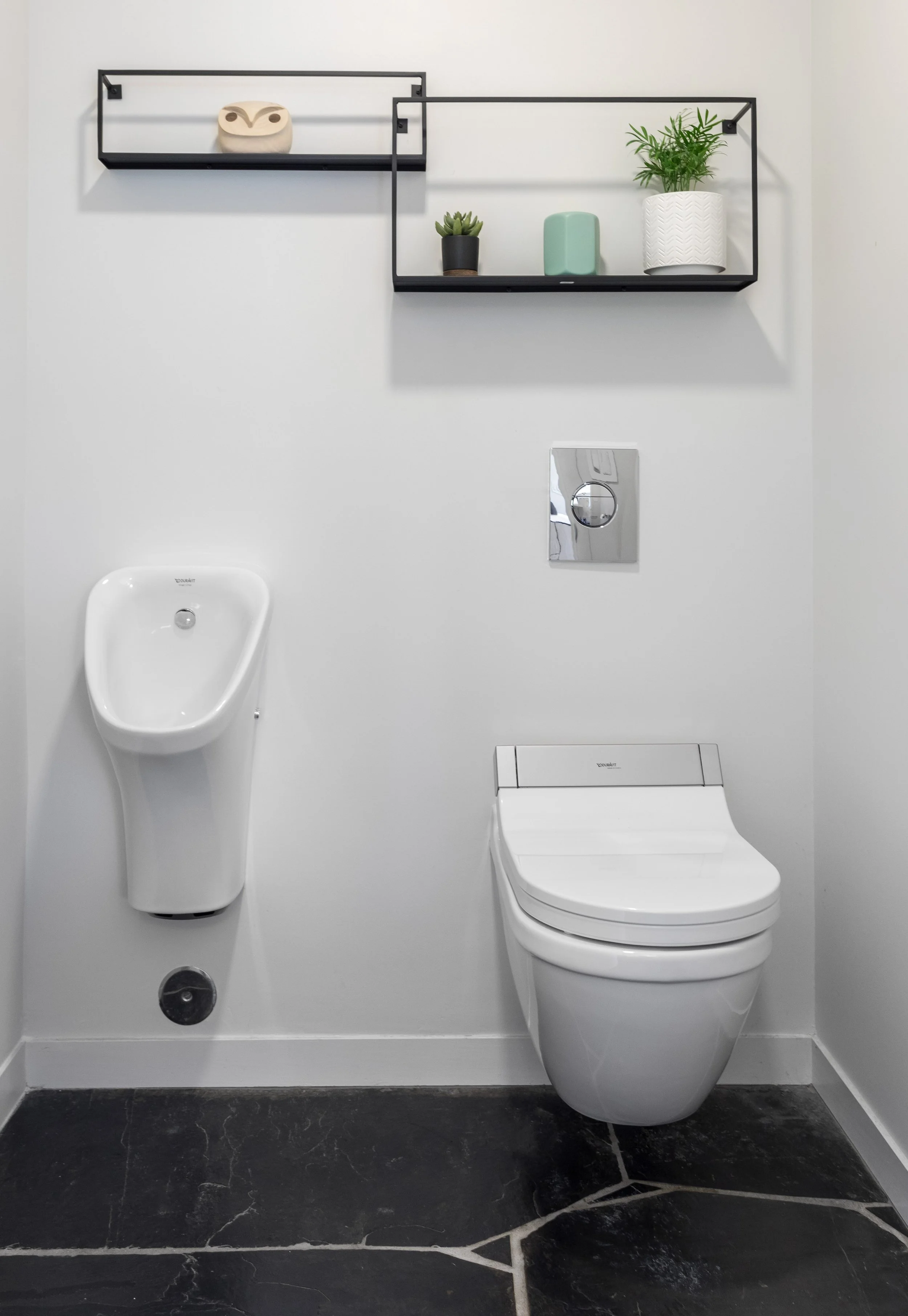 Modern bathroom with a wall-mounted toilet, a small wall-mounted urinal, a black metal shelf with decorative items, and a black marble floor.