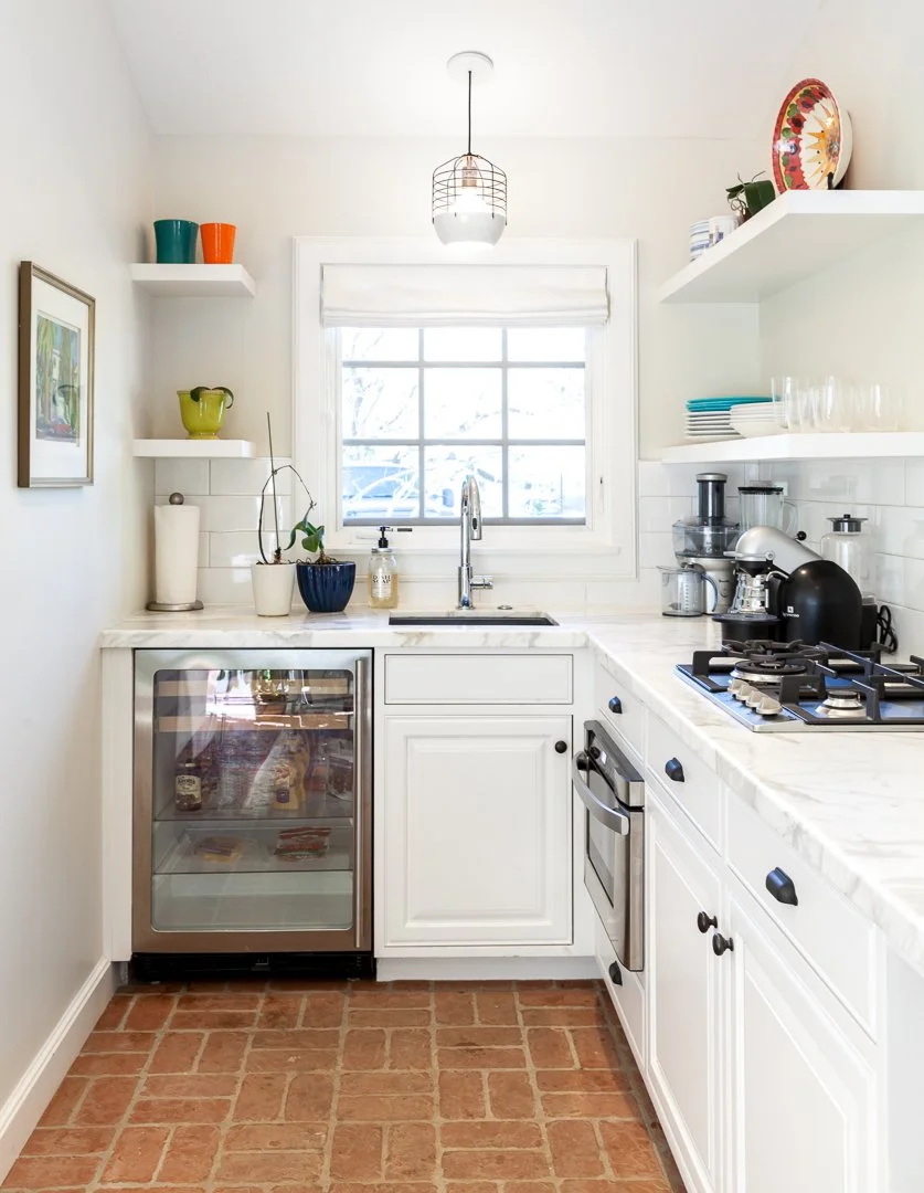 A cozy kitchen with white cabinets, open shelves, a window above the sink, and a brick floor. The counter has a small wine fridge, a sink, and various kitchen appliances, with colorful dishes and plants for decor.