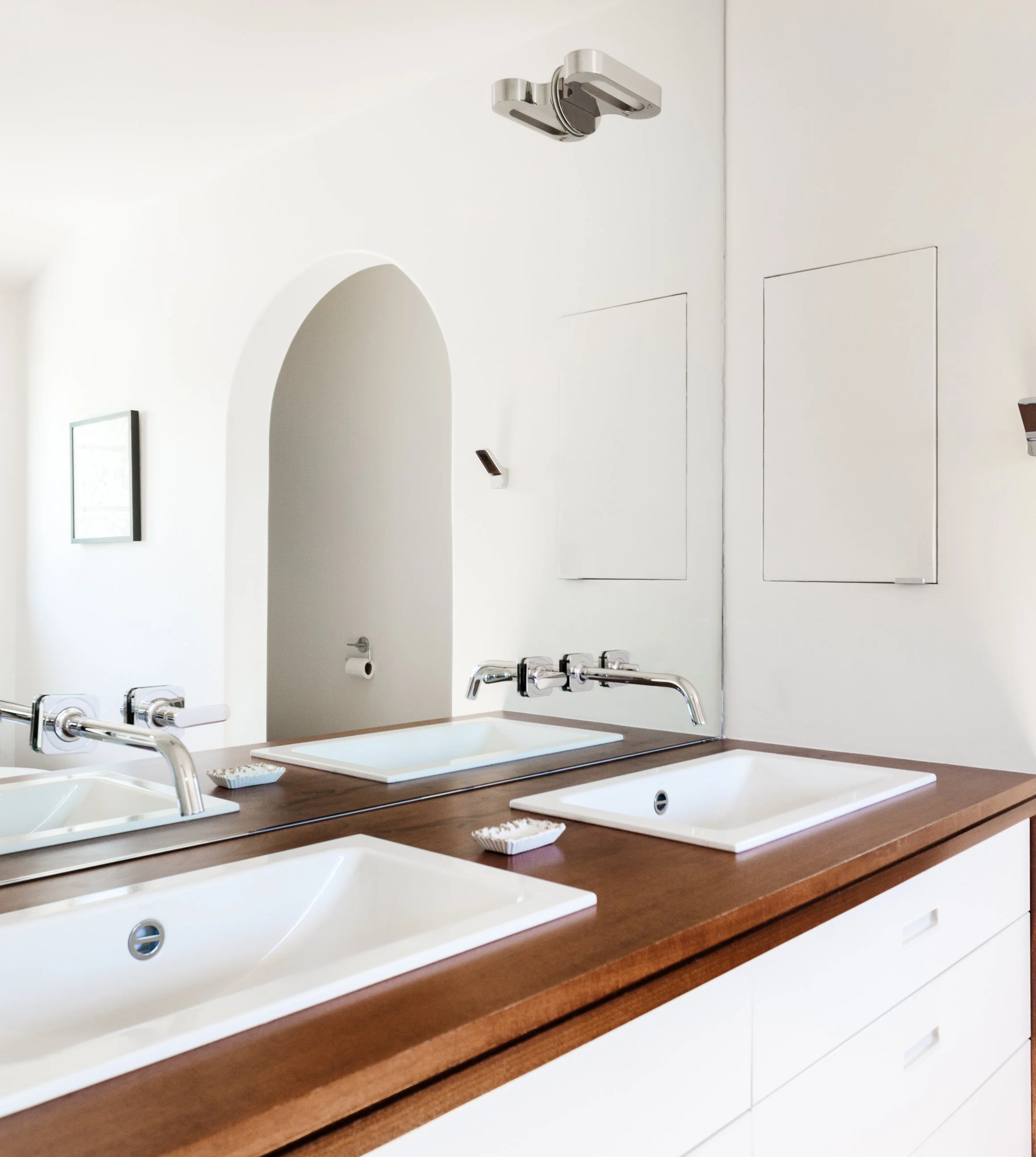 Bathroom double vanity with two white sinks and mirror. Wall-mounted faucets above each sink, and a soap dish on the wooden countertop. Minimalist decor, white wall with built-in cabinets, and a small wall-mounted light.