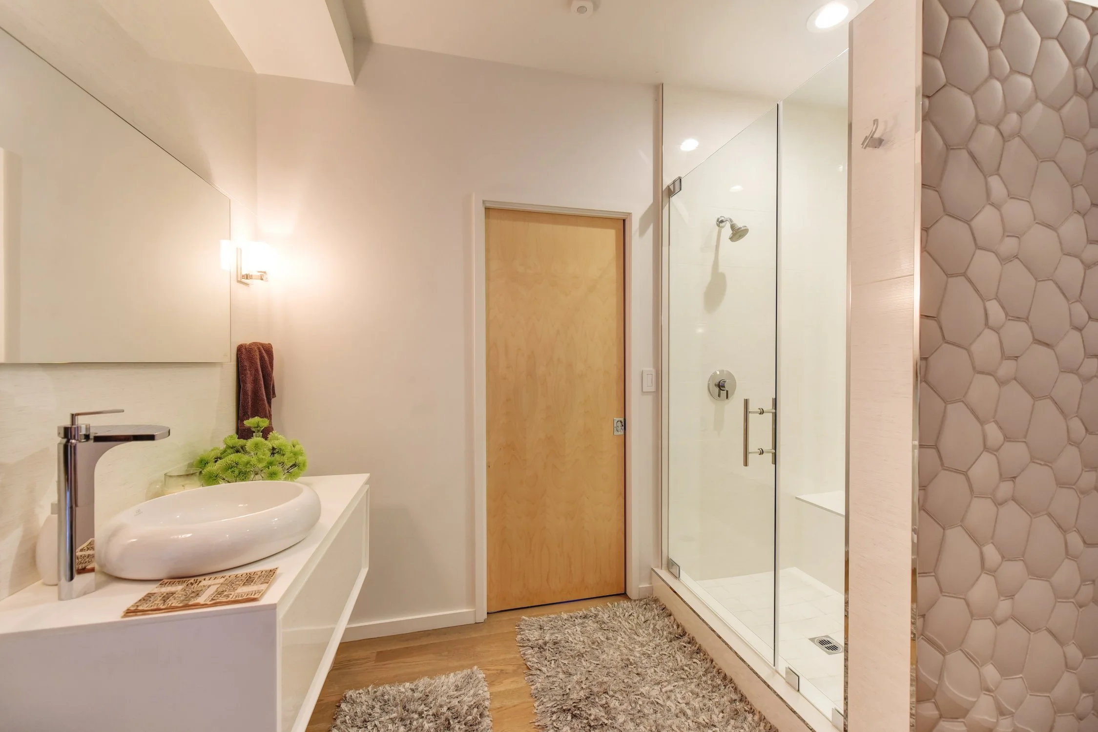 Modern bathroom with a white vanity, vessel sink, green plants, brown towel, acrylic shower with glass door, wooden door, wood flooring, and textured hexagonal wall tiles.