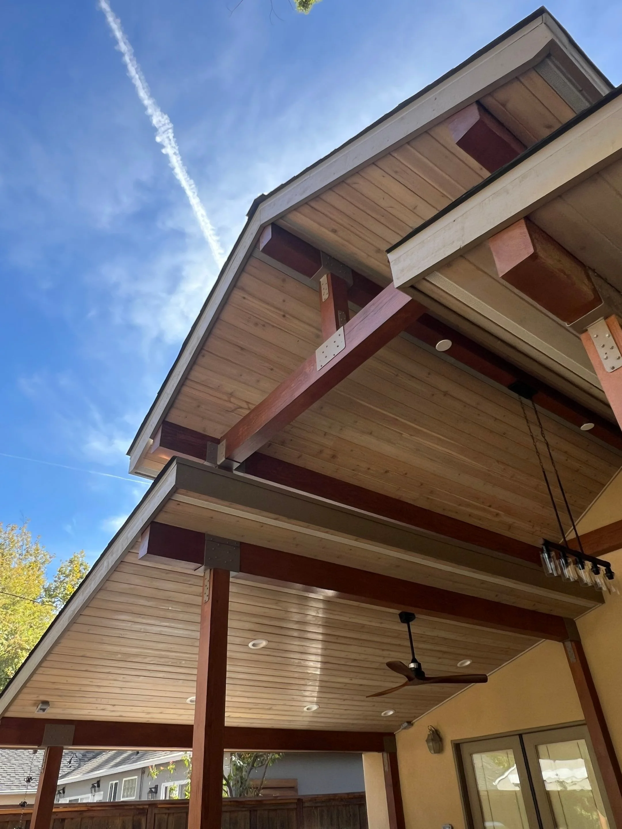 View of a modern house's ceiling and roof eaves from below, showing wooden panels, support beams, a ceiling fan, and a hanging light fixture, with a blue sky and trees in the background.
