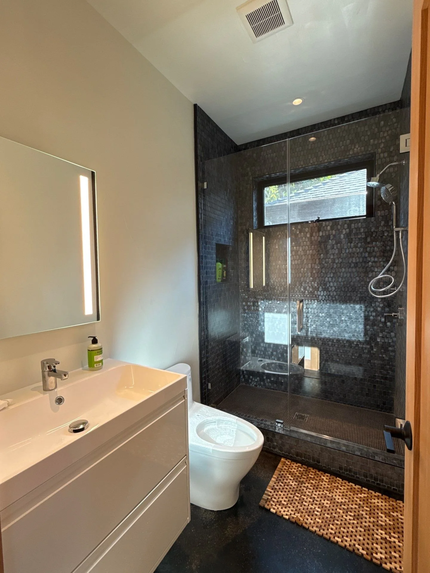 Modern bathroom with beige wall, white vanity with a mirror above, a white toilet, and a black tiled walk-in shower with a glass door, a window, and a rain shower head. There is a wooden door partially open on the right and a brown patterned bathroom