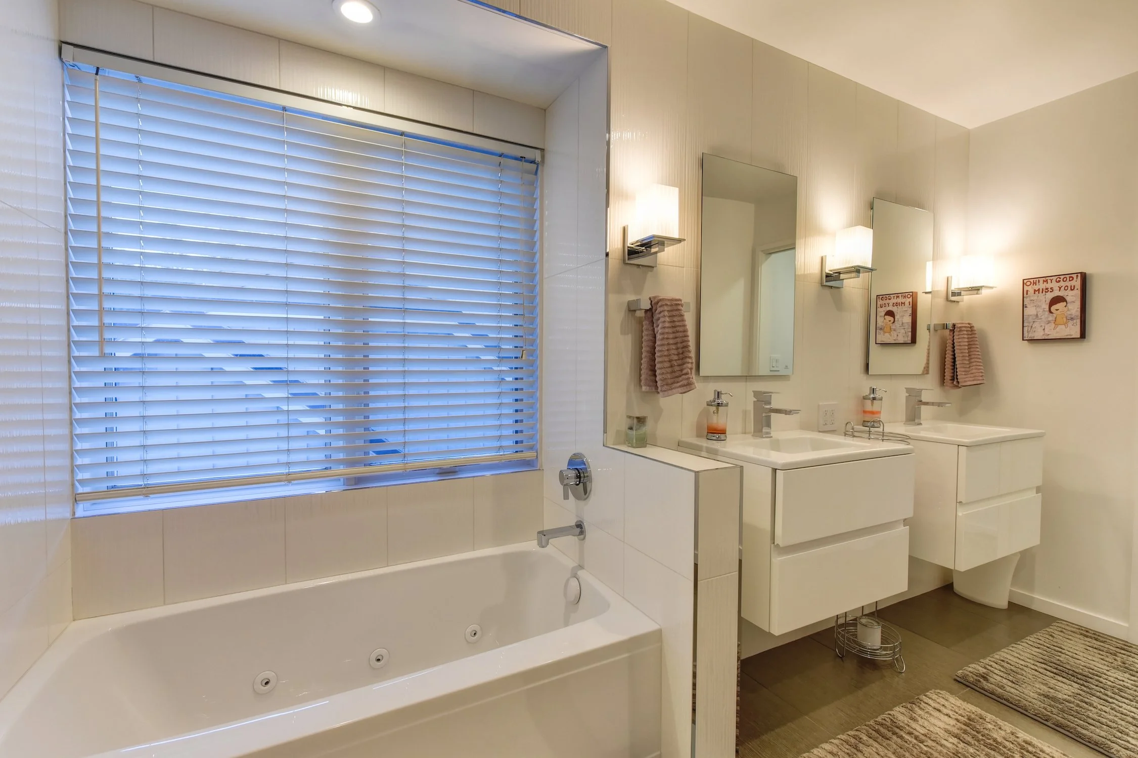 Modern bathroom with a large window with white blinds, a white bathtub, a double sink vanity with mirrors, and wall-mounted lights. There are two pink towels hanging and framed artwork on the wall.