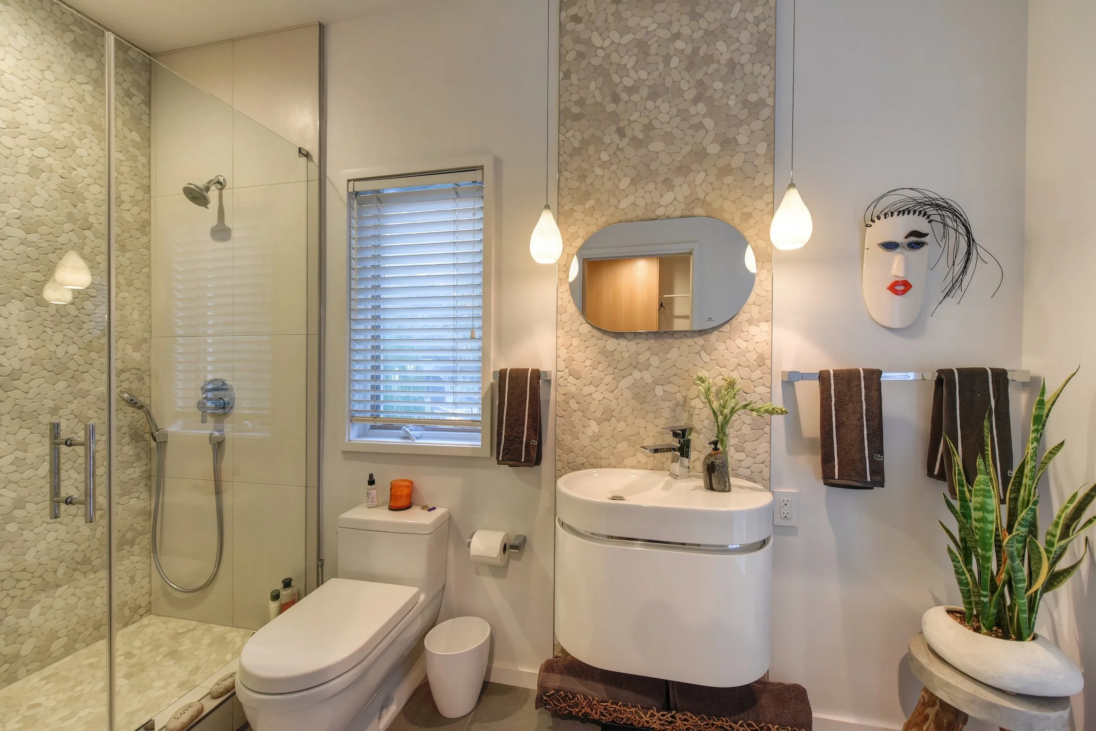Modern bathroom with a pebble-tile shower, white toilet, and a white curved vanity with a sink. Decorative mirror and wall art, warm lighting fixtures, window with blinds, and a potted plant.