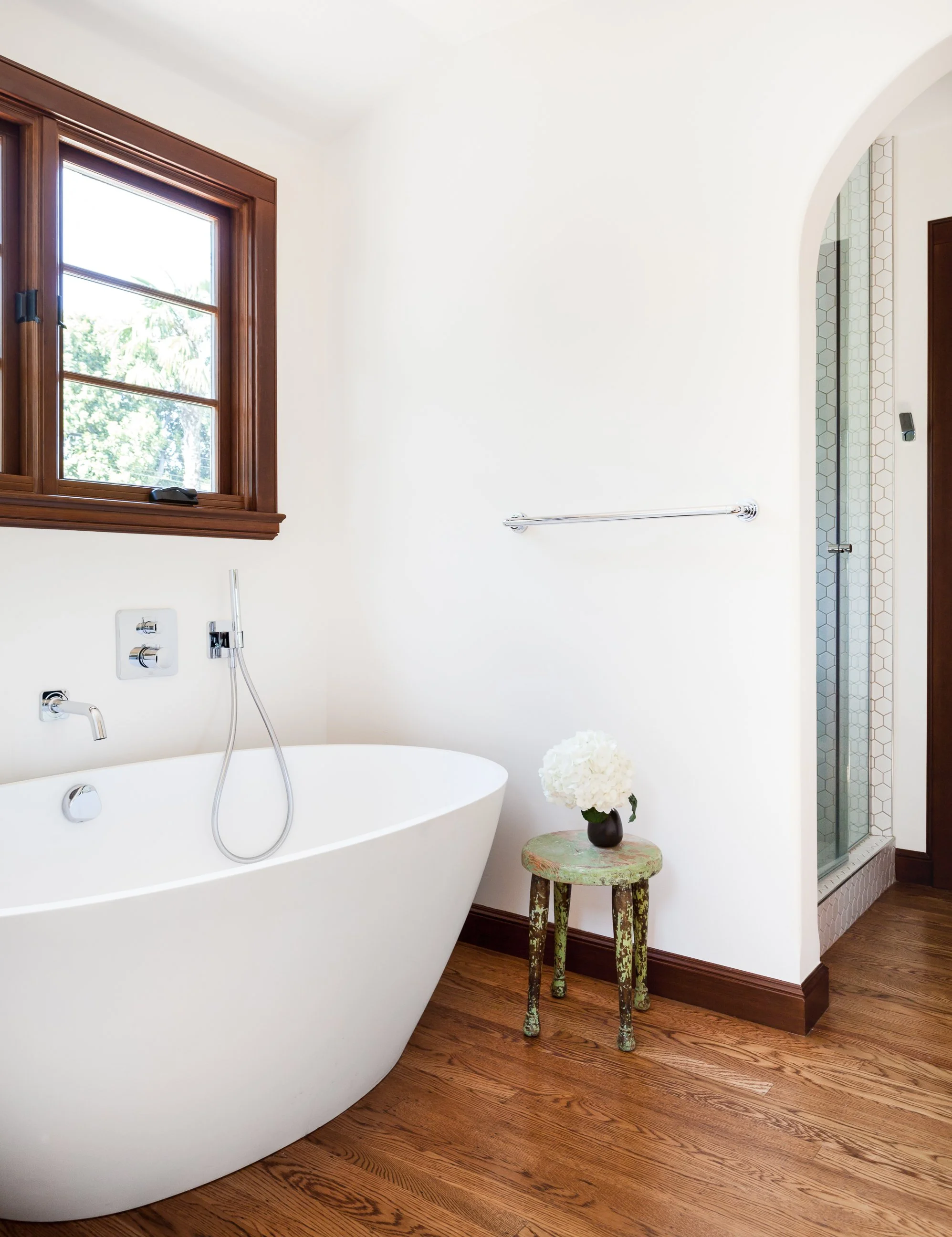 A modern bathroom with a freestanding bathtub, a wooden-framed window, a small rustic stool with a flower vase, and a walk-in shower with hexagonal tiles.