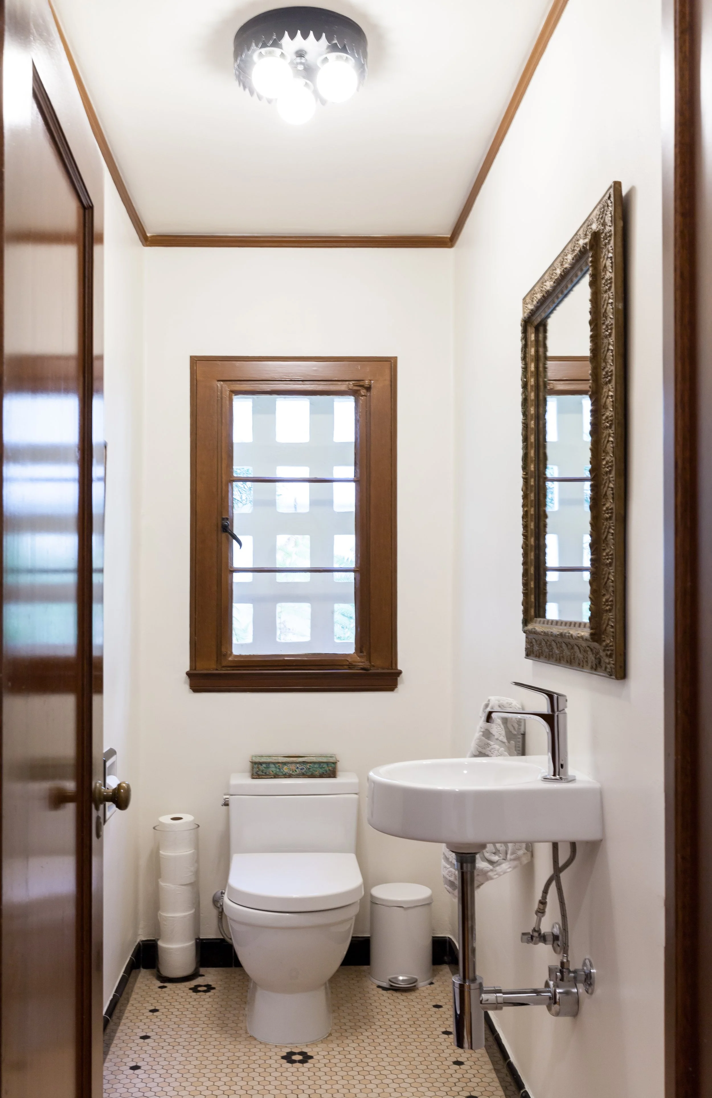 Small bathroom with a toilet, sink, and a window with wooden trim. Features a large framed mirror, a toilet paper roll, and a trash can. The ceiling has a modern light fixture.