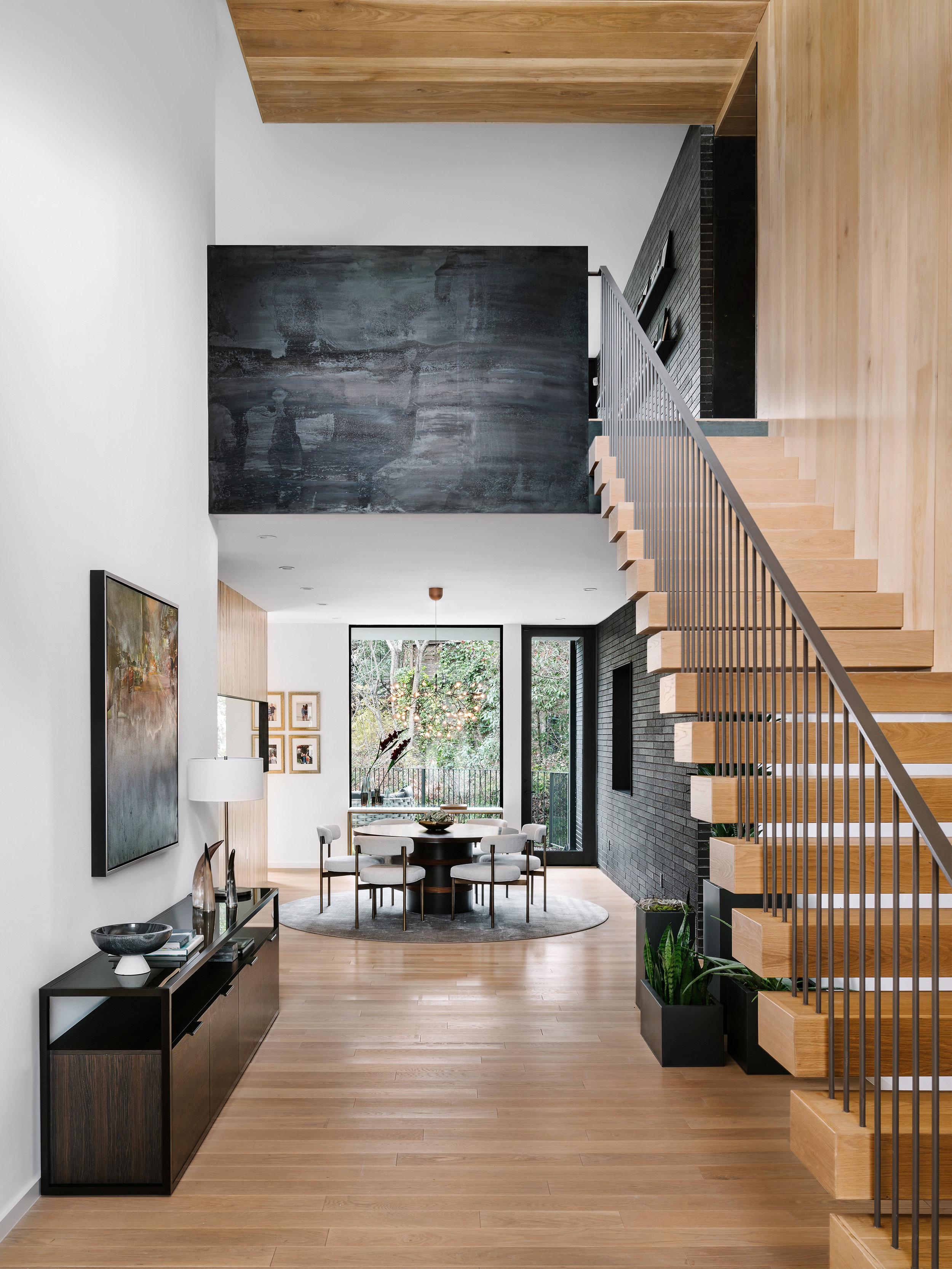 Modern, open-concept living area in the Pecos Residence with wooden staircase, dining set, console table, art, and large windows offering a forest view.