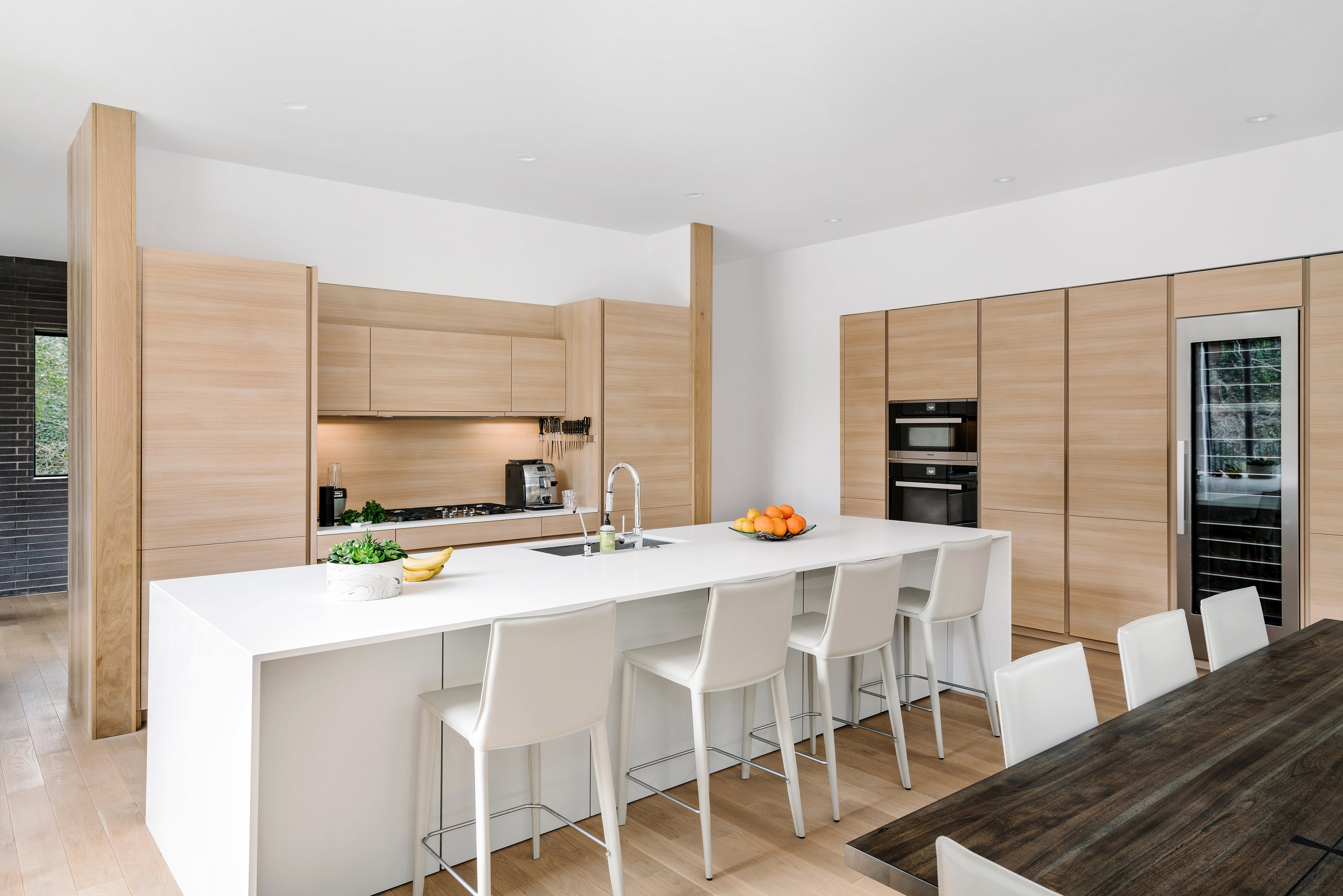 Modern kitchen in the Pecos Residence with wooden cabinets, white countertops, a large island with bar stools, and a dining table on the right.